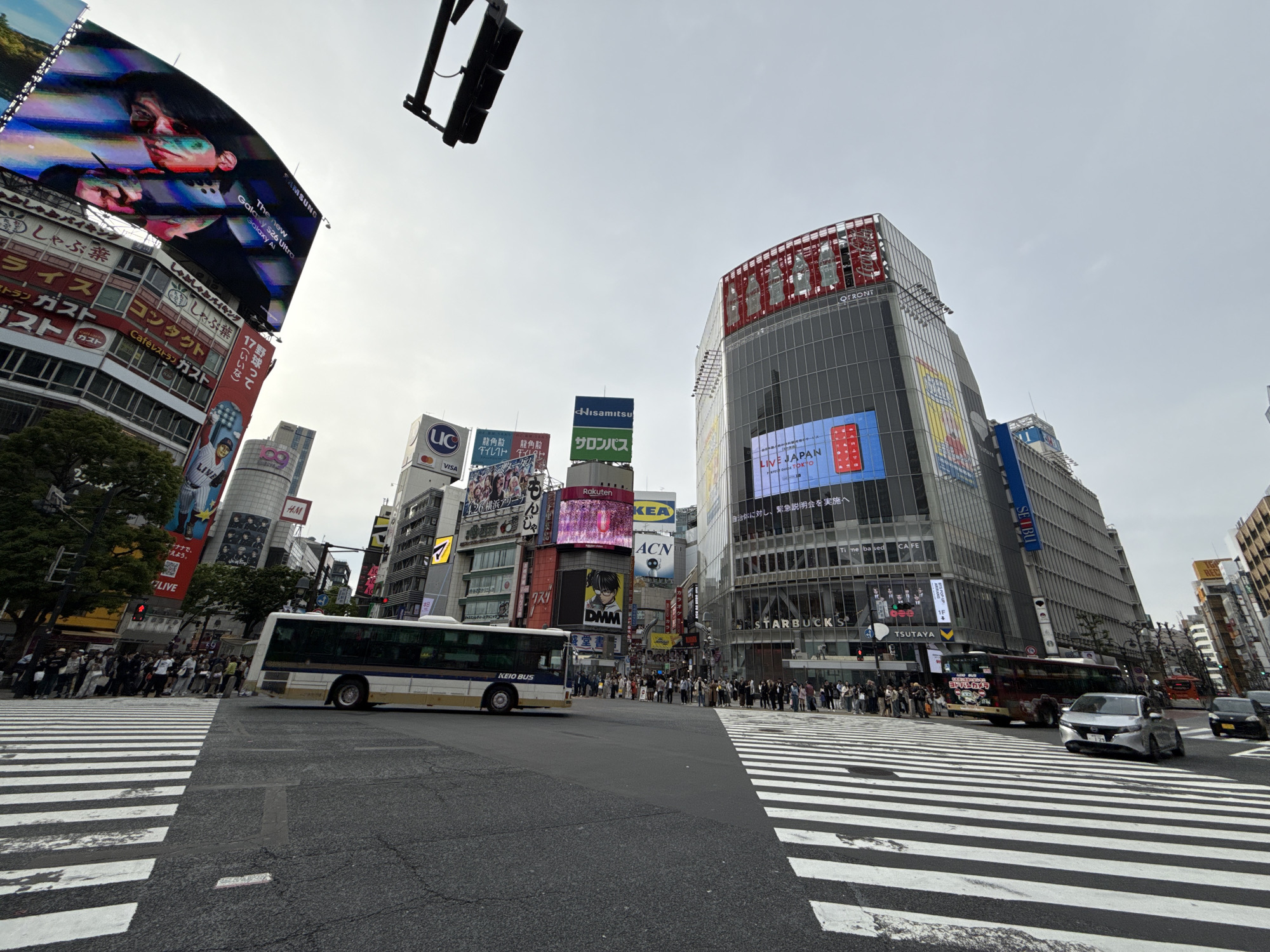The image captures a wide-angle view of a bustling urban intersection in Shibuya, Japan, under an overcast sky. Numerous pedestrians are gathered at the marked crosswalks, waiting for traffic to clear, while vehicles, including a prominent white and blue "KEIO BUS," navigate the intersection.
The scene is dominated by tall, modern buildings adorned with a multitude of large electronic billboards and static advertisements. Notable advertisements include a "SAMSUNG Galaxy S26 Ultra" ad featuring a person with a stylus, a "LIVE JAPAN" sign, "Rakuten," "IKEA," "DMM," "Hisamitsu サロンパス" (Salonpas), "UC VISA," and "H&M." One large building prominently displays "QFRONT" at its top, with "STARBUCKS" and "TSUTAYA" signs visible below. Other visible businesses include "SEIBU," "ヨドバシカメラ" (Yodobashi Camera), and various Japanese restaurants such as "しゃぶしゃぶ葉" (Shabu-shabu Yo) and "ガスト" (Gusto).
The activity depicts a typical busy daytime scenario in a major city, with people and traffic in motion or waiting at the crossing. The overall impression is one of dynamic urban life, characterized by dense infrastructure and widespread commercial advertising.