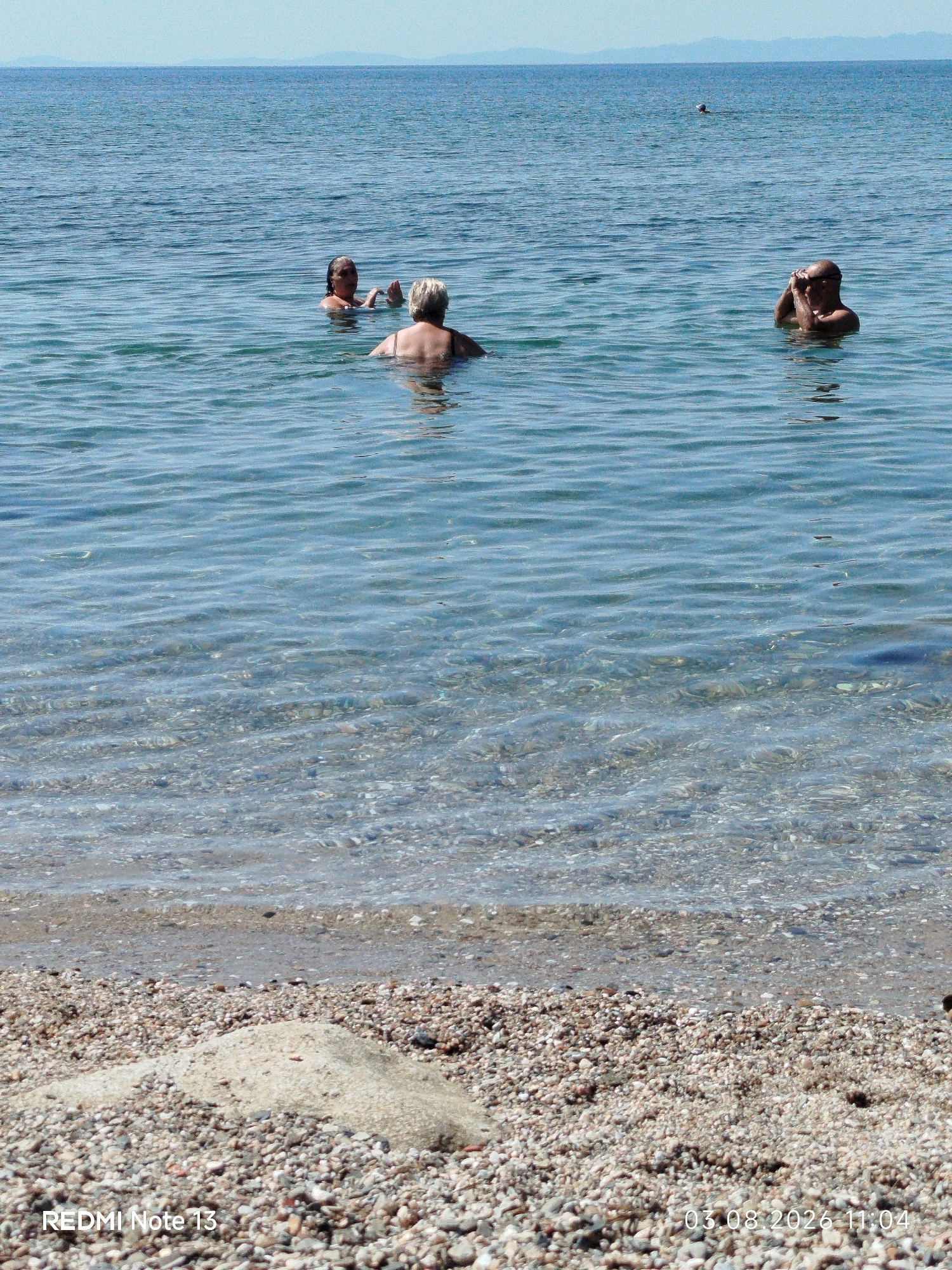 The image shows a clear blue sea with small waves, and a pebble beach in the foreground. There are four people visible in the water. In the mid-ground, two women are swimming, with one facing the camera and the other facing away. To the right, a man is partially submerged, holding his hands up to his face. Further in the background, a lone swimmer is visible. The sky is a pale blue, and distant hills can be seen on the horizon. The water is transparent enough to see the pebbles on the seabed. The timestamp on the image reads "03.08.2026 11:04" and "REDMI Note 13".