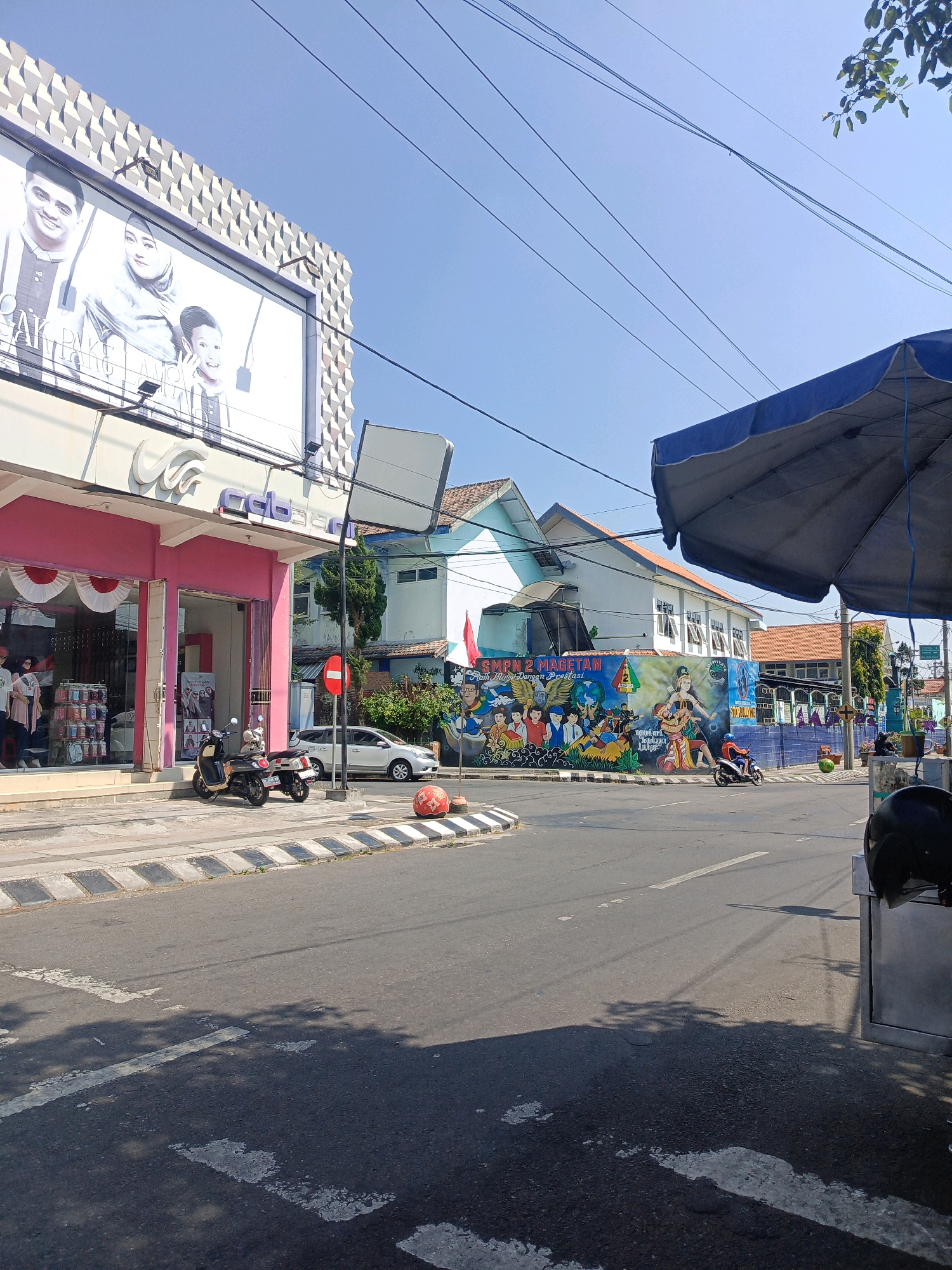 The image depicts a street scene in Sukowinangun, Indonesia. On the left, a pink building with large windows displays mannequins and merchandise. Above the storefront, a billboard shows images of three people. A sign with white text on a blue background is visible on the building. Two scooters are parked on the sidewalk in front of the pink building. Across the street, a white car is parked. Further down the street, a building is adorned with a colorful mural depicting people and the text "SMPN 2 MAGETAN". Several utility lines run overhead. A blue umbrella is visible in the foreground on the right. The ground is paved asphalt with white markings. The sky is clear and blue.