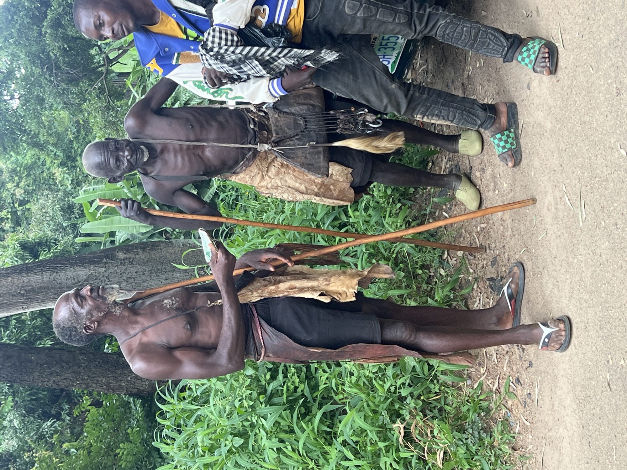 Three men are standing outdoors in a rural setting, with lush green foliage behind them and a dirt path in the foreground. A portion of a vehicle is visible on the right.
The man on the left is shirtless, wearing black shorts and a traditional leather skirt or loincloth, and sandals. He has a thin build and is holding a piece of paper or money in his right hand, and a wooden staff in his left.
The man in the middle is also shirtless and appears older, wearing a similar leather garment and what seem to be slip-on shoes. He is holding a wooden staff.
The man on the right is wearing a blue and yellow jacket over a yellow shirt, dark jeans with a textured pattern, and checkered sandals. He is holding a black and white checkered scarf.
The overall impression is a candid, possibly ethnographic, scene. The location is identified as the city of Jos, Nigeria.
