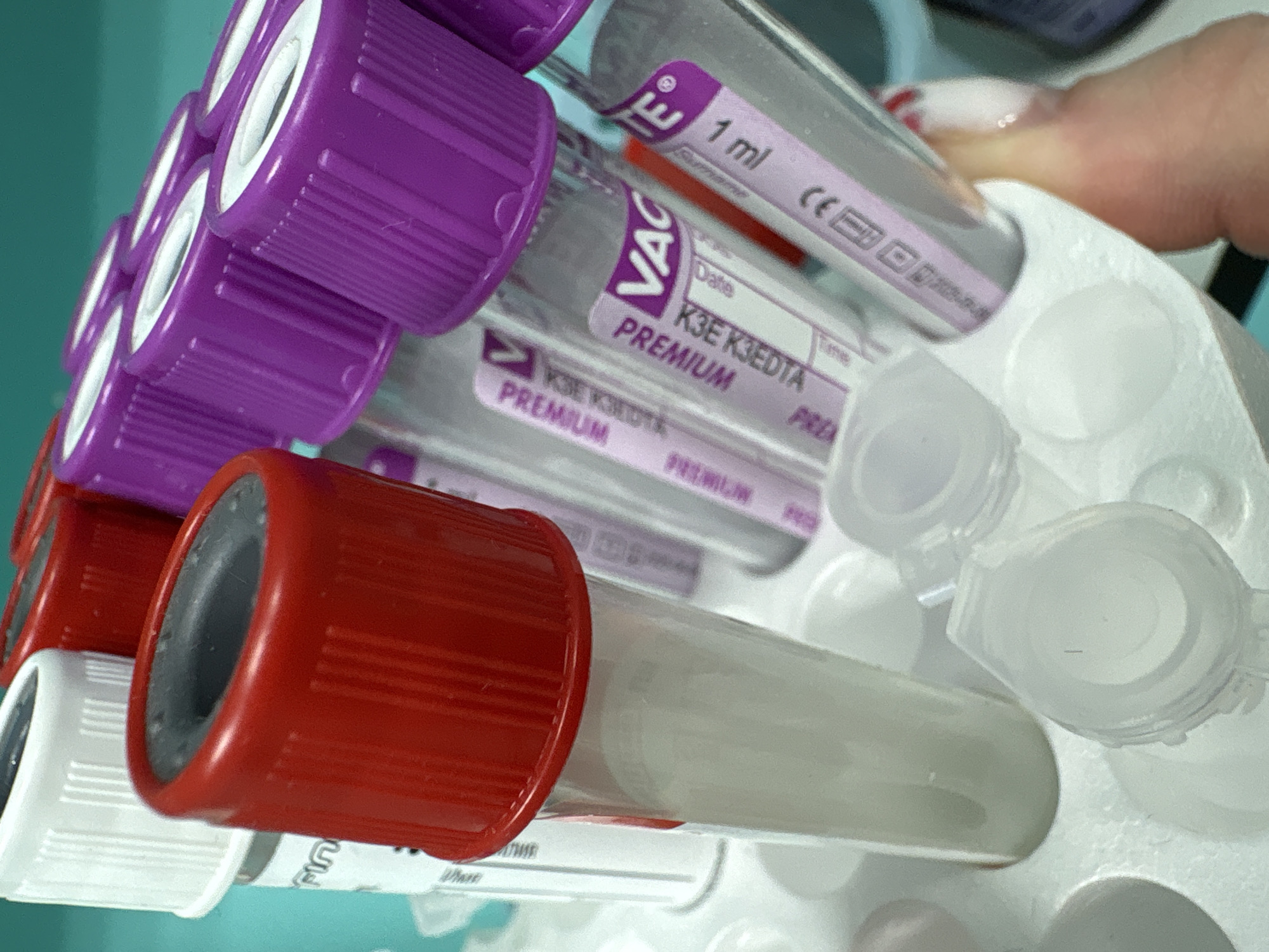 The image shows a close-up of several blood collection tubes held in a white plastic rack. The tubes are of varying types, indicated by the different colored stoppers: purple, red, and white. The purple-topped tubes are labeled "VACUETTE® PREMIUM K3E K3EDTA." The tubes appear to be unused, as they are empty or contain only a small amount of additive. The background is a light teal color. The location is not identifiable based on the image content. No people are visible in the main focus of the image, although there is a blurred area that might be a fingertip with nail polish visible at the right edge of the image. There are no discernible actions taking place.