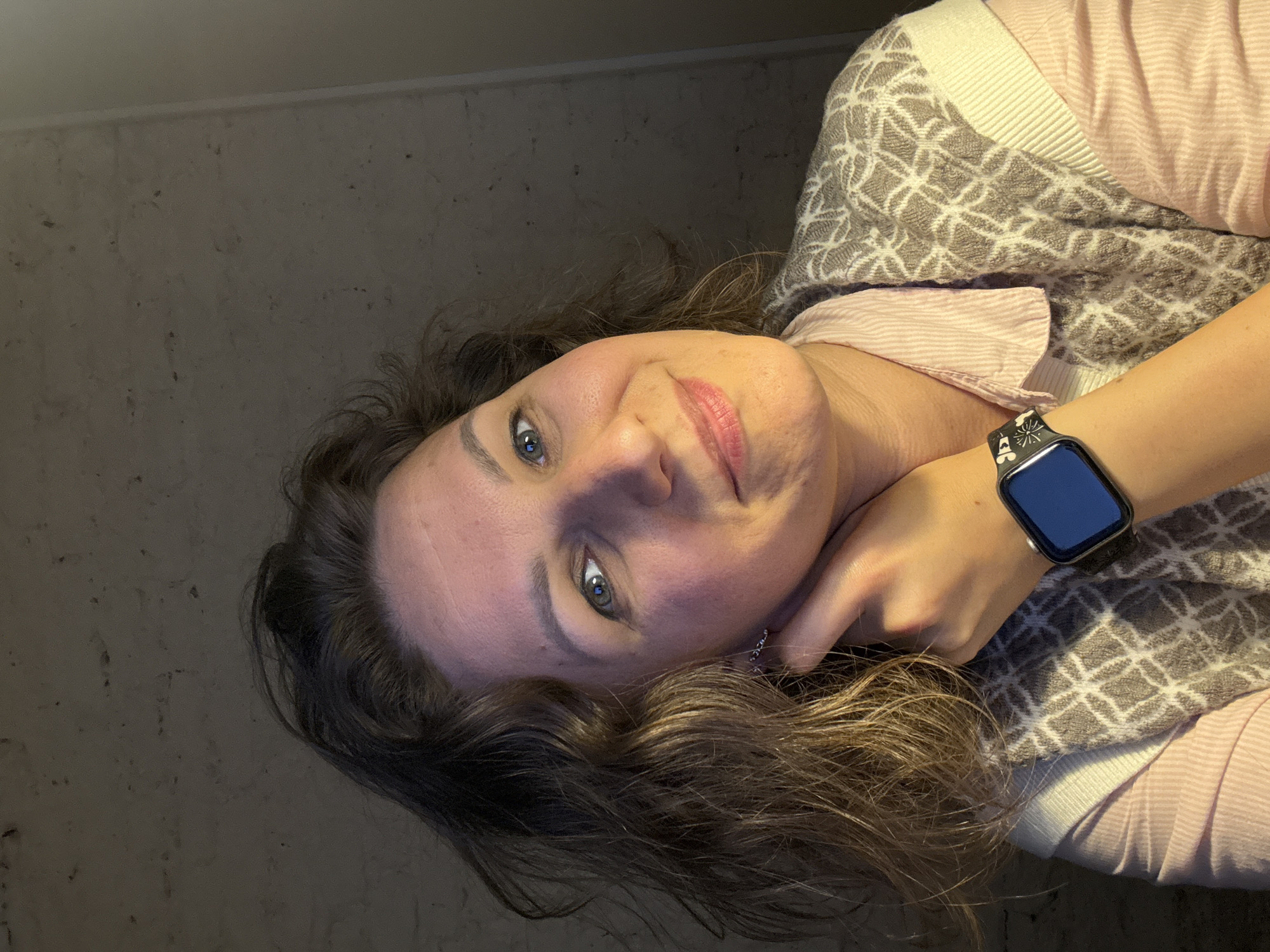 A woman with fair skin, blue eyes, and light brown curly hair is photographed indoors. She is wearing a pink collared shirt under a patterned cardigan in shades of white and grey. A smart watch with a dark face is visible on her left wrist. The background is a plain, dark wall. It is not possible to determine the location as Saint Petersburg, Russia.