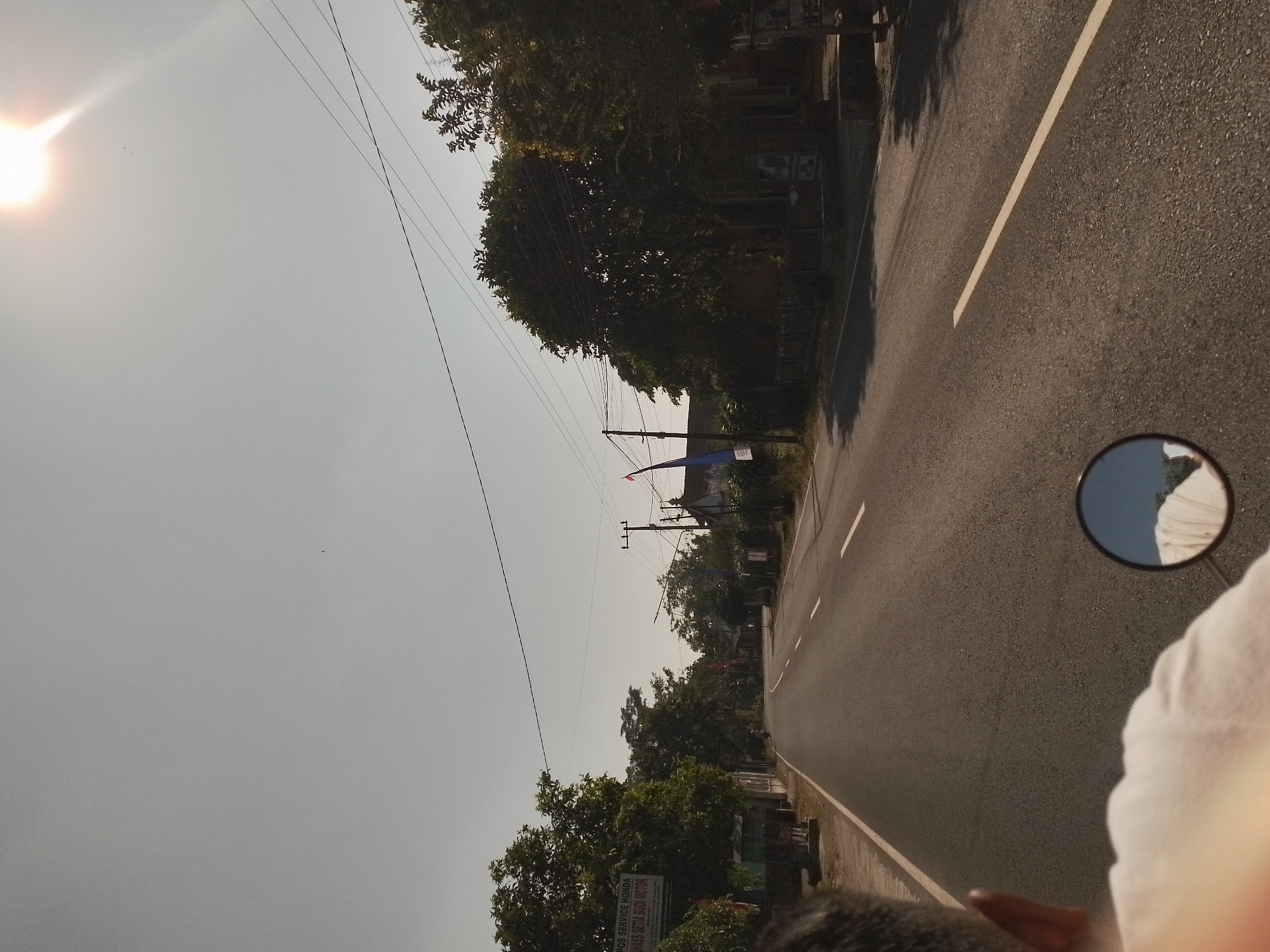The image depicts a road scene in Ngancar, Indonesia, under a bright, hazy sky with the sun visible in the upper right corner, casting lens flare. A person's head and shoulder, wearing a white shirt, are visible in the foreground from behind, suggesting the photo is taken from a moving vehicle, likely a motorcycle, as indicated by the presence of a round rearview mirror in the lower center. The road itself is paved and appears to be a two-lane street with white dashed lines marking the lanes. To the left of the road, a sign for "POS SERVICE HONDA HASS SETIA BUDI MOTOR" is visible, along with some foliage and buildings. The right side of the road features trees, more buildings, and electrical poles with overhead wires. Red and blue flags are also present on poles along the roadside. The overall environment suggests a rural or semi-urban setting.