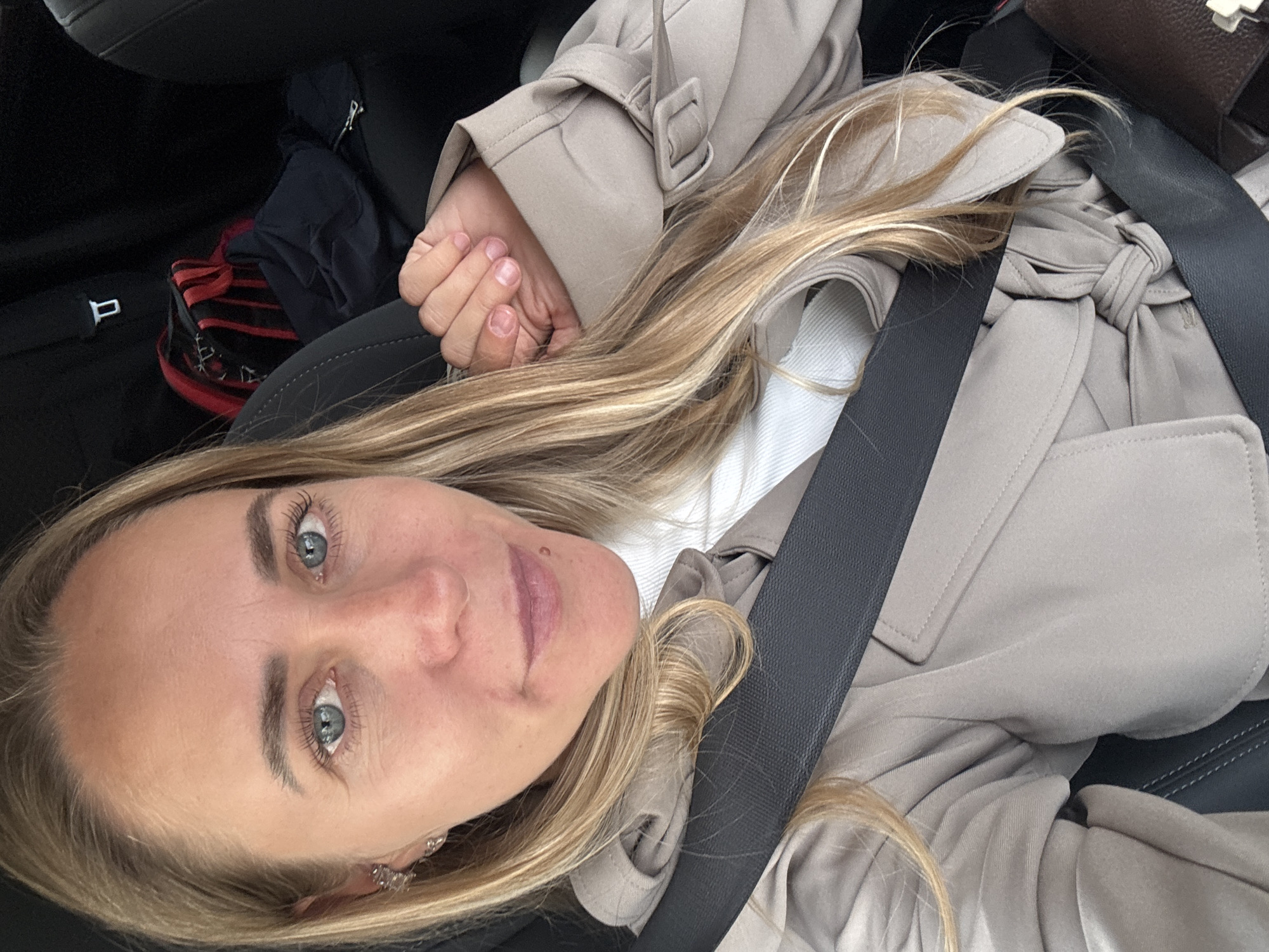 A blonde woman with blue eyes and fair skin is captured in a selfie from the chest up, seated in the passenger seat of a car. She is wearing a beige trench coat over a white turtleneck. A black seatbelt is fastened across her body. In the background, on the back seat, a red and black striped bag and a dark blue garment are visible. The car's interior, including a black seatback, is also visible. The lighting suggests it is daytime. The image does not contain any specific information that would allow for its location to be determined as Chaikovsky, Russia.