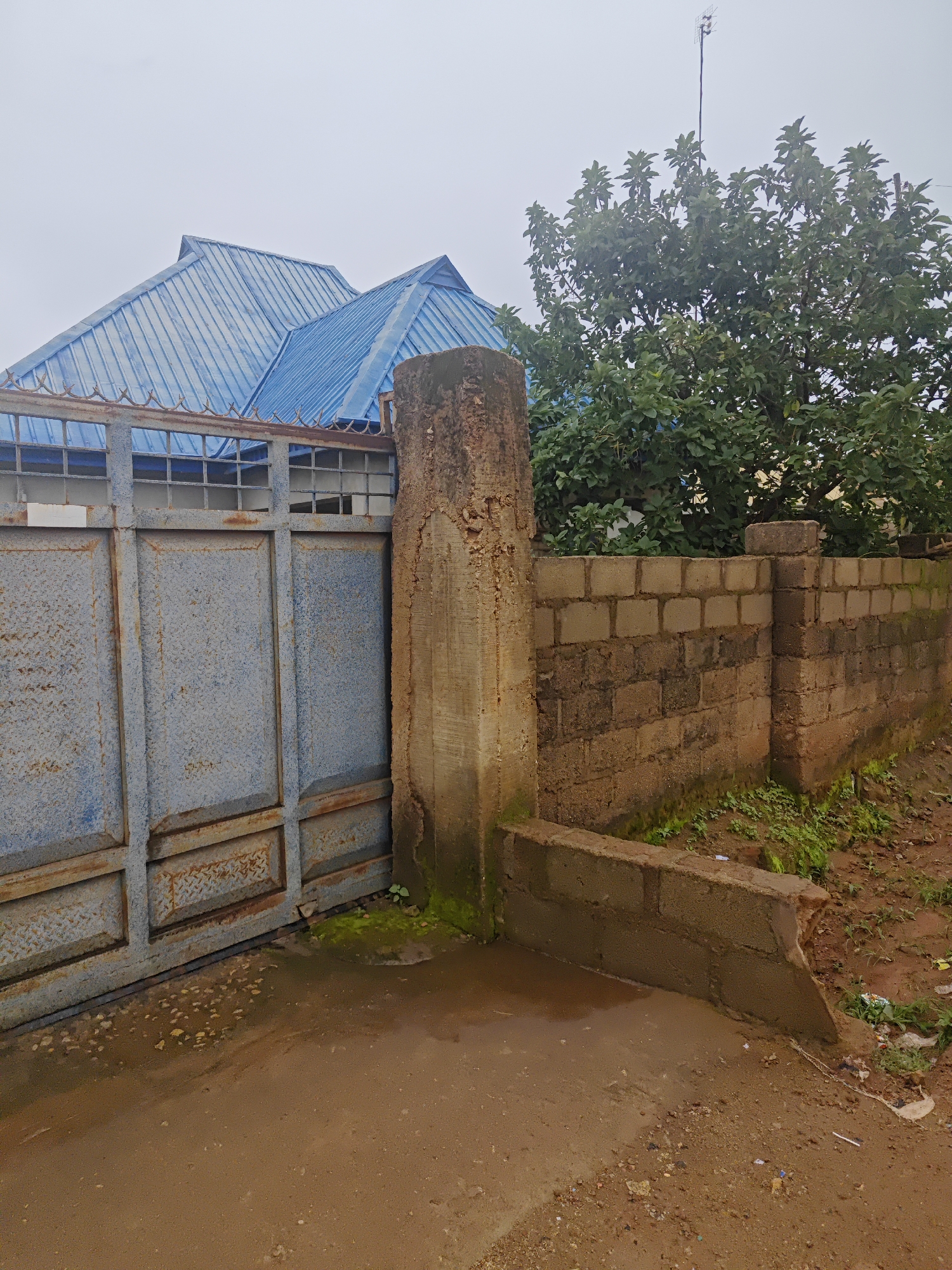 A weathered, blue metal gate, showing signs of rust and damage, stands partially open in front of a building with a blue metal roof. To the right of the gate is a thick, rough concrete pillar and a low, cinder block wall. Behind the wall, lush green foliage from a tree is visible against an overcast sky. The ground is a mixture of dirt and scattered debris. The scene appears to be in an outdoor, possibly residential area, within the city of Bukuru, Nigeria.