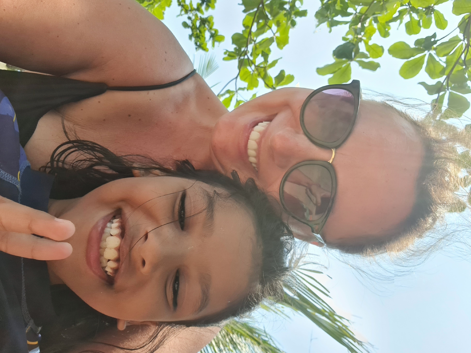 A mother and daughter take a selfie while outside. The mother is wearing sunglasses and a black spaghetti-strap top. The daughter is wearing a blue shirt and making a peace sign with her hand. They both are smiling and their teeth are visible. The background shows green leaves and a blue sky. The location is outdoors in Đà Nẵng, Vietnam.
