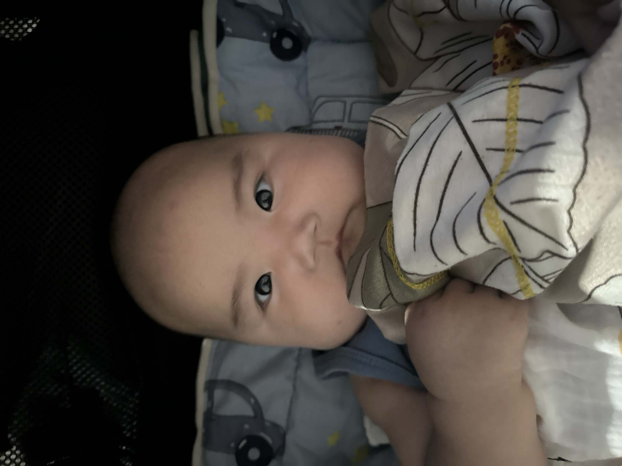 A close-up shot shows a baby, likely a boy, looking directly at the camera. The baby has a round face, dark eyes, and is bald. He is wearing a light blue sleeveless shirt. The baby is wrapped in a patterned blanket with black lines and yellow accents. The background appears to be a stroller or crib, with a black mesh fabric visible at the top and a light blue, patterned lining with images of elephants and stars. The overall lighting is dim, with the face of the baby well-lit. The scene is indoors.