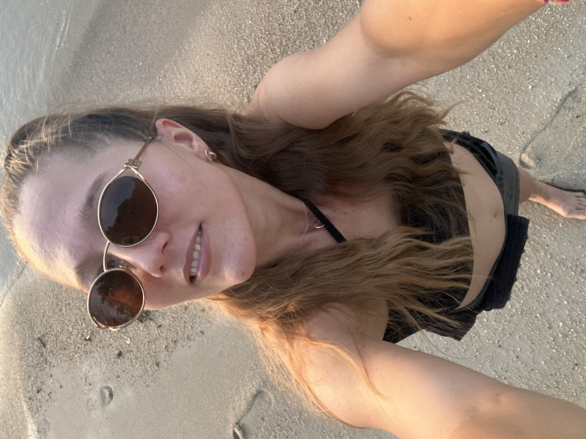 A young woman with long, wavy blonde hair is taking a selfie on a sandy beach. She is wearing sunglasses and a black top. The sun is low in the sky, casting a warm glow on the scene. The ocean is visible in the background. The location is the beach in Baan Thong Po, Thailand.