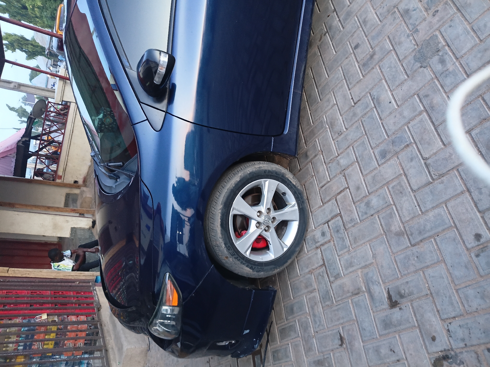 The image captures a street scene in Jalingo, Nigeria, with a dark blue sedan parked on a brick-paved surface. A person wearing a black cap and a colorful t-shirt is seated against a wall to the left of the vehicle, near a storefront displaying various bottled beverages behind a metal grate. The car's front wheel is visible, featuring a silver rim and red brake calipers. The environment appears to be an urban setting, with architectural structures and foliage visible in the background. The lighting suggests it might be late afternoon or early evening.