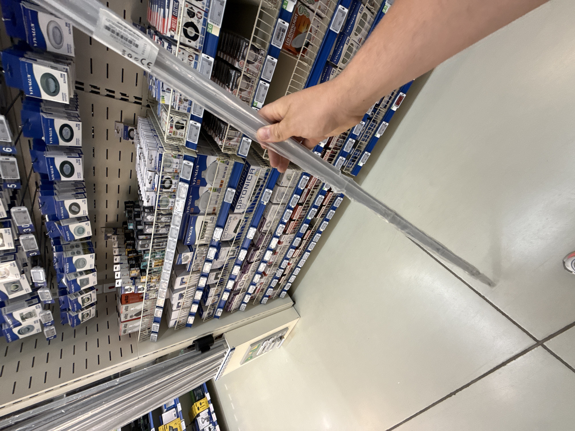 The image captures a view of a retail store aisle, specifically in a section dedicated to lighting fixtures and related products, located in Stara Zagora, Bulgaria. The shelves are stocked with various boxes of lighting, including spotlights, LED lights, and downlights. A person's hand is prominently featured in the foreground, holding a long, clear plastic-wrapped bar, likely a lighting component or accessory. The aisle flooring is a light-colored, tiled surface with dark grout lines forming a grid pattern. The overall impression is one of a well-organized retail environment, possibly an electronics or home improvement store.