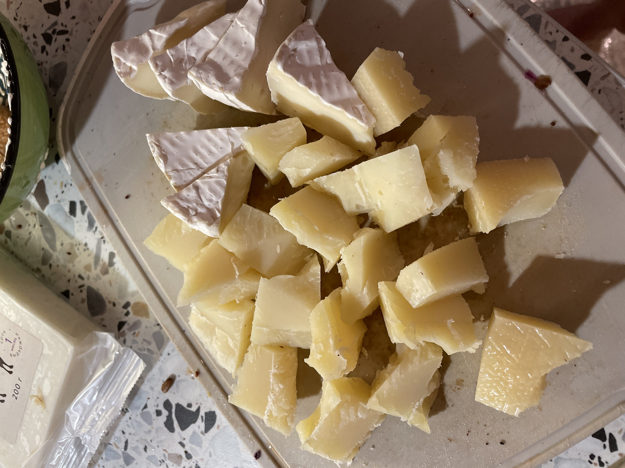 A close-up, eye-level shot captures a collection of cut cheeses arranged on a light gray cutting board. The cheeses are varied, with some appearing to be hard, aged varieties cut into irregular, angular chunks, and others being softer, triangular wedges of brie-style cheese with a white rind. The cutting board has a textured, speckled surface. In the upper left corner, a portion of a cheese package is visible, showing text and an illustration of an animal. A green bowl with what appears to be nuts or other food items is partially visible in the upper right background. The lighting is somewhat dim, creating soft shadows.