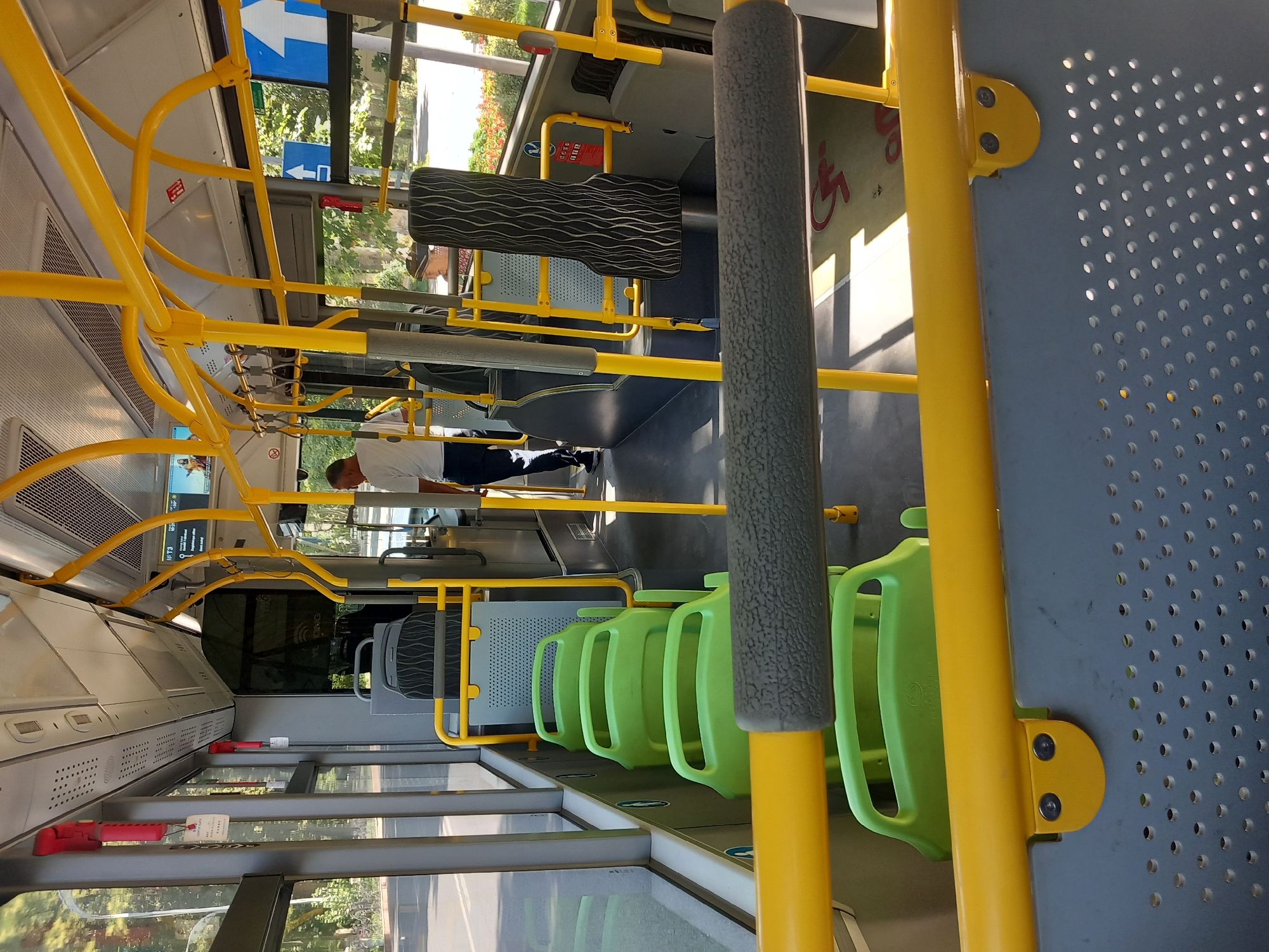 An interior view of a bus, likely in Samarkand City, Uzbekistan, shows a row of empty, light green plastic seats on the left side. A man in a white t-shirt and dark pants stands in the aisle on the right, facing away from the camera. Yellow handrails and support poles are prominent throughout the bus. The floor has a pattern of light and shadow, with a red wheelchair symbol visible in the lower right corner. The environment outside the bus windows appears to be sunny with trees and buildings visible.