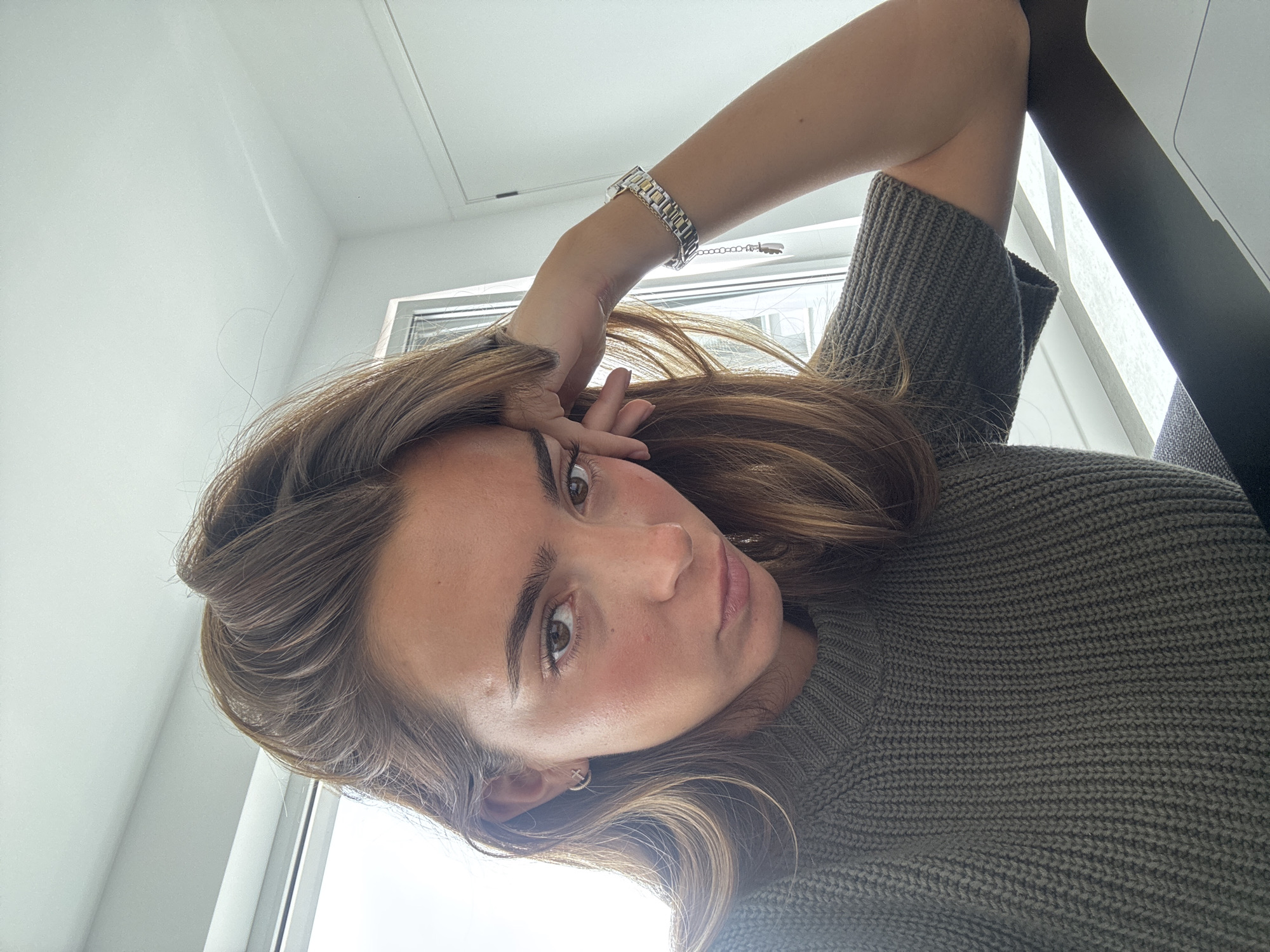 The image is a bright, close-up portrait of a young woman, likely a selfie, taken indoors with ample natural light. She has long, wavy, medium-brown hair that falls around her shoulders, dark eyebrows, and expressive brown eyes, looking directly at the camera with a calm and neutral expression. Her head is tilted, and her left arm is raised, with her hand gently touching her temple.
She is wearing a thick, olive-green or dark grey-green knitted sweater with a prominent ribbed texture. On her left wrist, a two-tone metal watch with silver and gold-toned links is visible. A small, subtle earring, possibly a cross design, can be seen in her right ear.
The background features bright white walls and a large window or glass pane, suggesting a daytime setting with clear natural illumination. A dark, undefined object, possibly a laptop or furniture, is partially visible on the right side of the frame, casting a shadow against the wall. The overall scene conveys a relaxed and intimate moment. There is no visible text in the image.