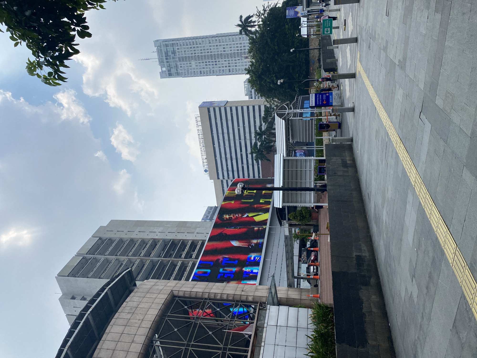 In South Jakarta, Indonesia, on a bright, cloudy day, a wide-angle shot captures a street scene. In the foreground, a paved sidewalk with tactile paving runs parallel to a dark, stone-tiled wall. Beyond the wall, a street is lined with cars and orange traffic cones.
Dominating the mid-ground is a large, illuminated digital billboard displaying colorful images and text, with the words "YOU GOT THIS" prominently featured. To the left of the billboard, a building with large windows is visible, showing reflections of the digital display. Behind the billboard, a multi-story building with rows of balconies stretches upwards. Further in the background, a tall, modern skyscraper stands against the sky, and another, slightly shorter building with horizontal windowed sections is visible.
A lush green tree with dark leaves partially obscures the top right of the frame. The sky is a pale blue, dotted with white clouds, suggesting a slightly hazy atmosphere. Several people are present in the scene, walking on the sidewalk or near the buildings, but their details are indistinct. Signage for "SHOPPING MALL" and "OFFICE TOWER" are visible near the entrance of one of the buildings. The overall impression is of a bustling urban environment.