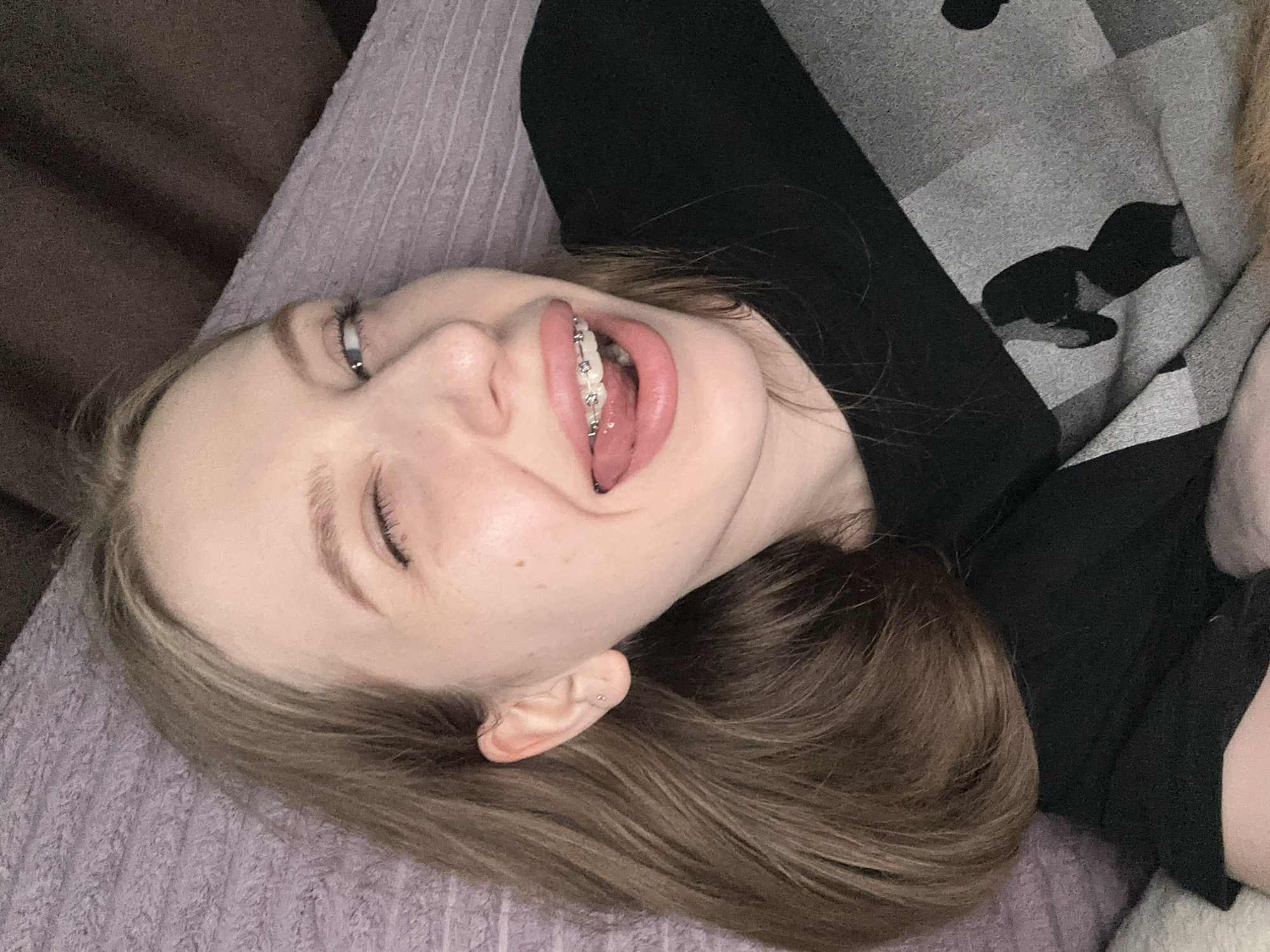 A young individual, likely female, is captured in a close-up, eye-level shot, lying down in a relaxed, indoor setting. The image is tilted, showing the person's head turned to their left, looking playfully towards the camera with a wide smile or laugh. Their mouth is open, revealing a slightly extended tongue and clear or white braces on the upper teeth. One eye is open, looking upwards, while the other is partially closed, conveying a joyful or mischievous expression.
The person has light brown hair, which is spread around their head, and fair skin. They are wearing a black long-sleeved top. A small, silver-colored stud earring is visible on their left earlobe. The background features a textured, light purple fabric beneath their head, and a darker, plain brown fabric in the upper left. On the right side of the frame, near their torso, there is a grey and black patterned fabric, possibly a blanket or another garment, displaying an abstract, blotchy design. The lighting is soft and indoor. There is no visible text.