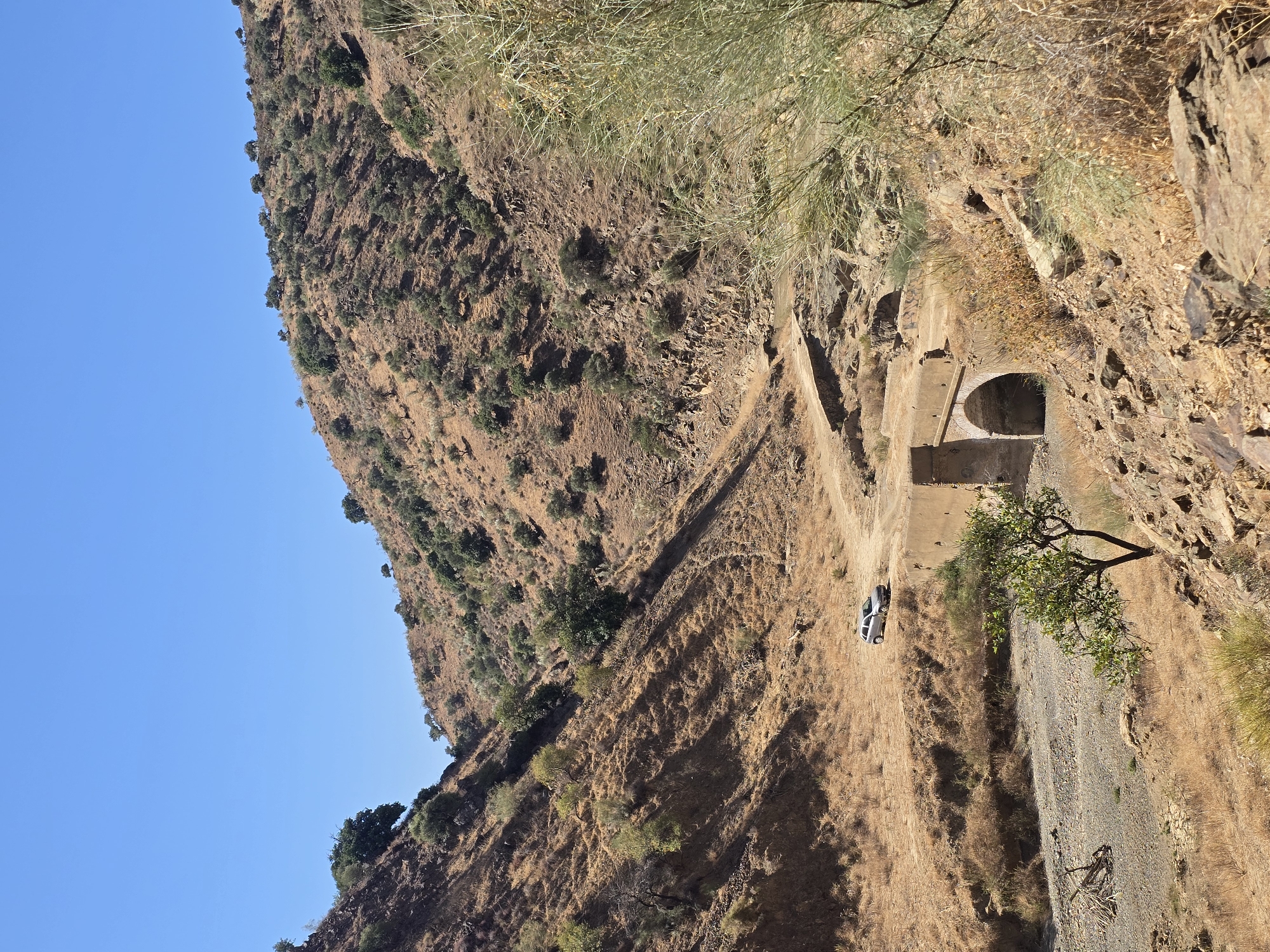 This is a photograph taken in the area of Almogía, Spain, depicting a dry, rocky, and arid landscape dominated by a steep hillside covered in sparse vegetation. A narrow, unpaved road winds its way through the valley, which also contains a dry riverbed. In the mid-ground, a small, arched concrete bridge spans the dry riverbed, with a silver SUV parked on the road near its edge. A lone tree with green foliage stands in the foreground to the left of the bridge. The sky is a clear, bright blue, indicating a sunny day. The overall impression is one of a remote, rugged, and dry environment.