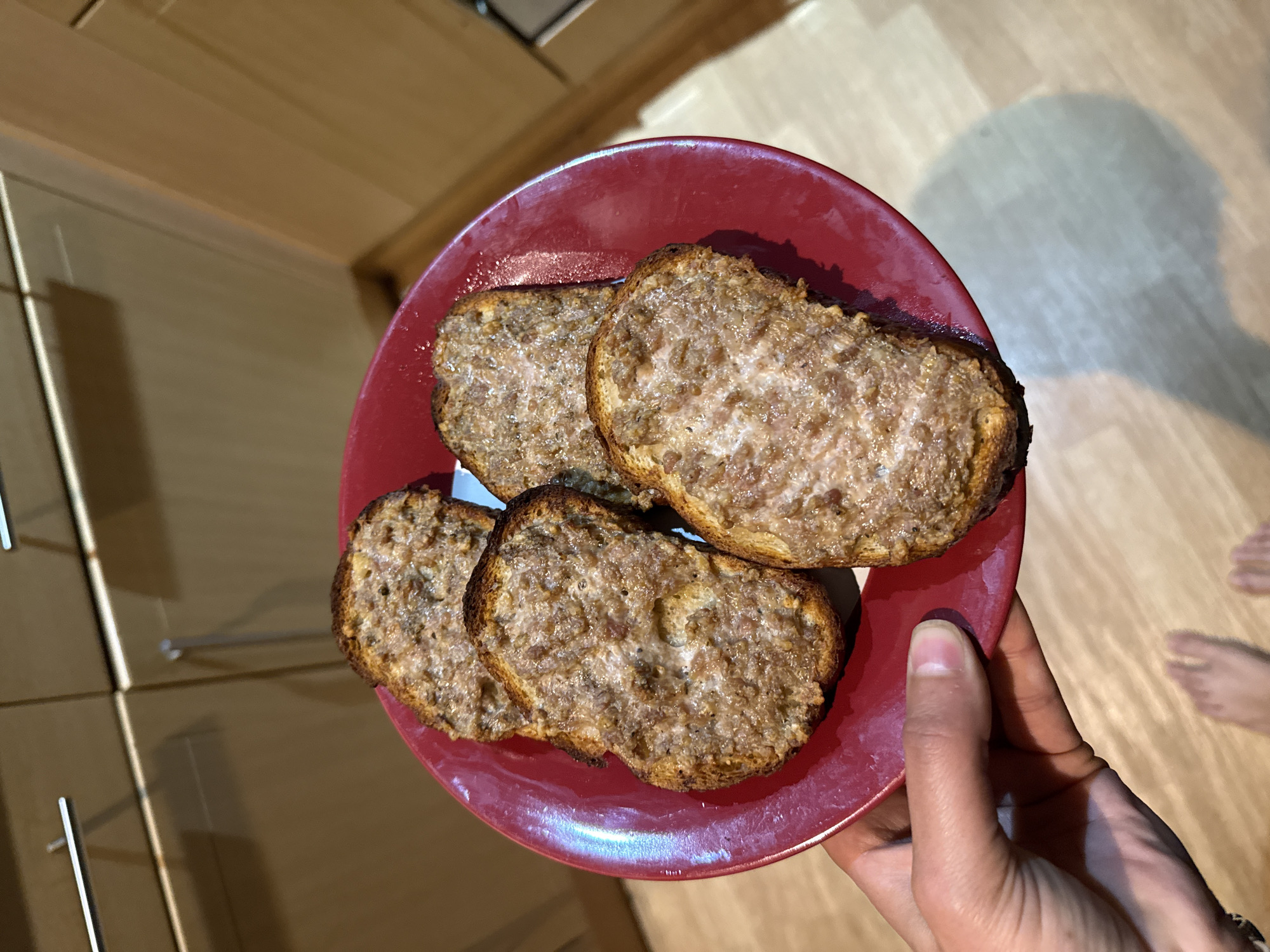 A person's hand, seen from a high angle, holds a red circular plate with four pieces of food. The food items appear to be toasted bread slices, rectangular or oval in shape, with dark brown, slightly burnt edges. Each slice is topped with a light brown, textured spread that looks somewhat granular or meaty, possibly a form of pâté or sausage meat spread, and appears moist or slightly oily.
The background shows an indoor setting, likely a kitchen or dining area, characterized by light brown wooden cabinetry or drawers on the left side, featuring silver horizontal handles. The floor beneath is made of light-colored wooden planks. A distinct shadow of the hand and plate is cast on the wooden floor to the right. Faint, blurred outlines or shadows of feet are also visible on the floor in the upper right background. The lighting suggests an indoor artificial source or ambient daylight. No text is visible in the image. The scene captures a moment of someone holding prepared food in a home environment.
