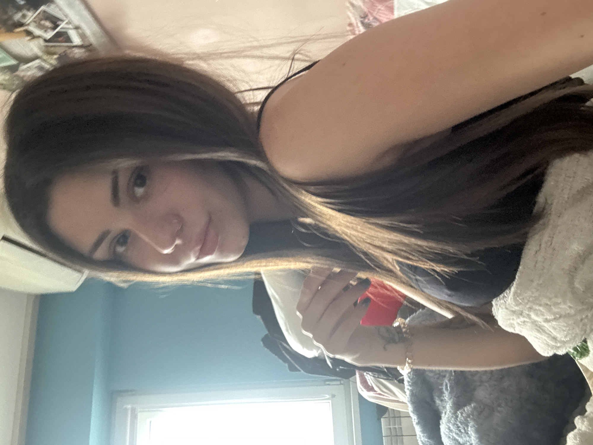 A woman with long brown hair is facing the camera. She is wearing a black top. Her right arm is bent, with her hand holding an object that is partially obscured. She has a bracelet on her wrist. The background includes a light blue wall with a window, and a portion of a bed with a blanket. The location is not discernible from the provided image.