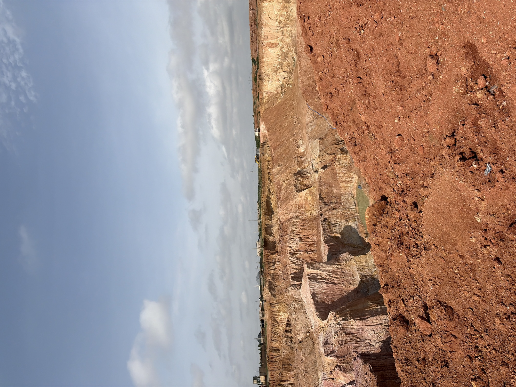 The image depicts an aerial view of an expansive open-pit mine, likely located in Kwang, Nigeria, characterized by its striking reddish-orange soil. The mine features sheer, layered cliffs of earth, sculpted by excavation activities, revealing strata of varying hues and textures, from pale yellow to deep rust. In the foreground, a large pile of loose, reddish soil and rocks dominates, with some scattered debris visible.
The midground showcases the intricate excavation patterns within the pit, with visible terraces and steep drops. A small, greenish pool of water is present at the bottom of the pit, suggesting accumulation from rainfall or groundwater. The distant background reveals the rim of the mine, where a collection of buildings and infrastructure can be observed, including what appears to be residential structures, utility poles, and possibly some industrial equipment. Two trucks are visible on the far edge of the mine, suggesting ongoing operations. The sky above is a pale blue, dotted with scattered white clouds, indicating fair weather conditions. There are no people visible in the image.