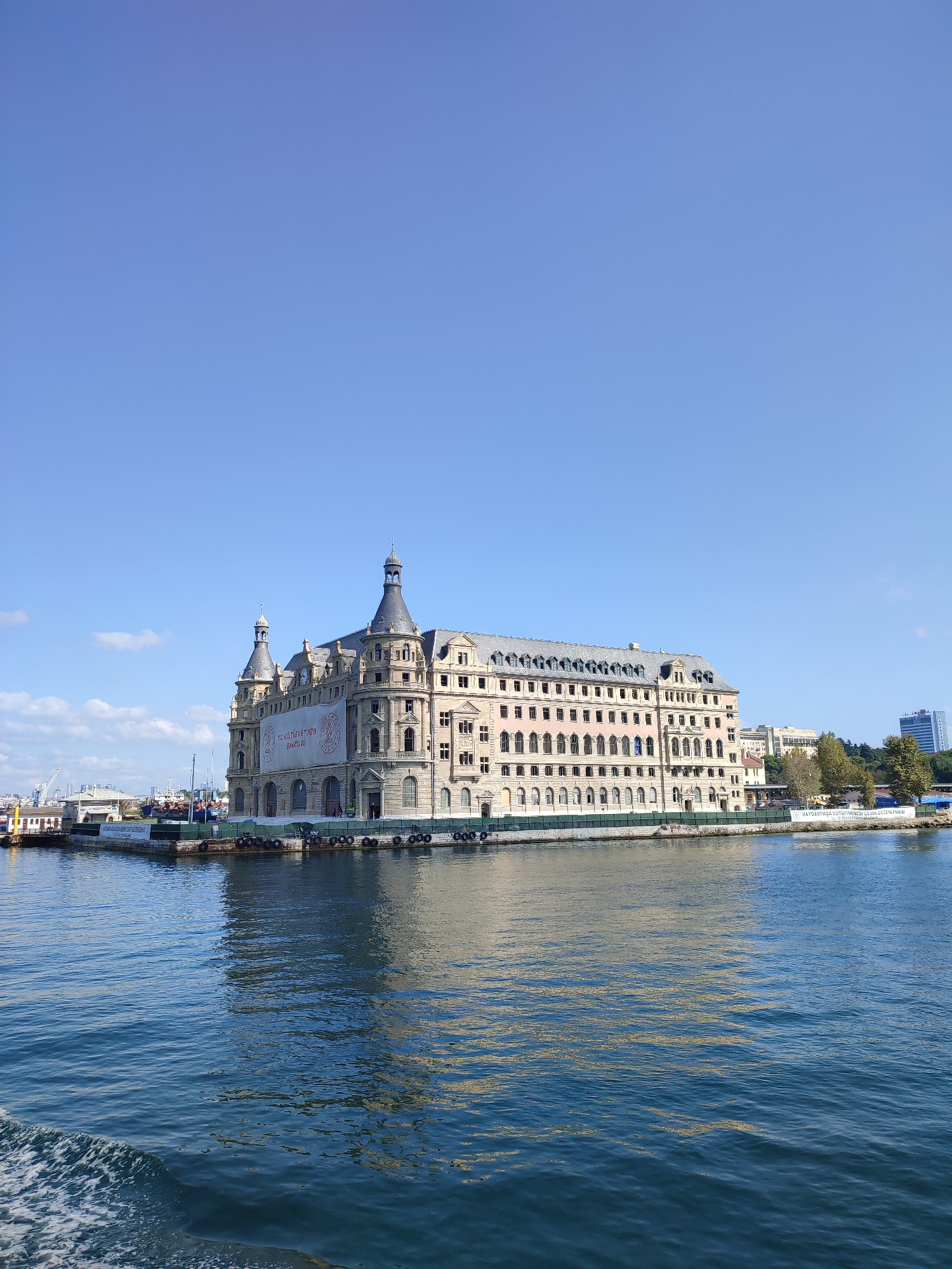 The image depicts Haydarpaşa train station, a prominent landmark in Kadıköy, Istanbul, Turkey. The grand, historic building, characterized by its ornate architecture and distinctive twin towers, stands prominently on the waterfront. It is illuminated by clear daylight under a bright blue sky with scattered white clouds. The building is reflected in the calm, blue waters of the Bosphorus Strait in the foreground. Several boats and ships are visible docked or sailing in the distance, suggesting a bustling port area. The overall scene conveys a sense of grandeur and historical significance.
