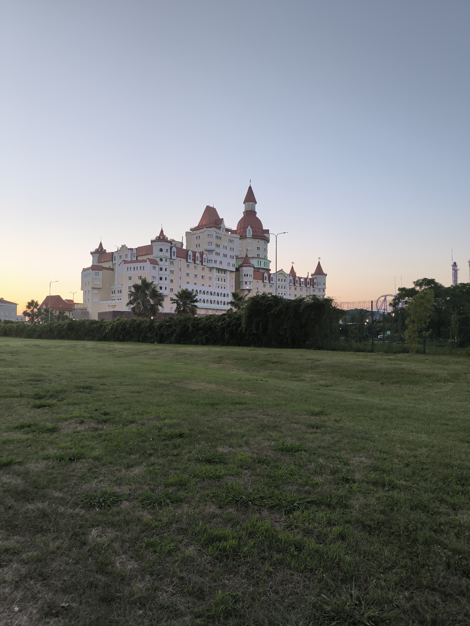 An exterior, eye-level, medium shot captures a brightly colored, castle-like hotel building on the left side of the frame, bathed in the soft light of dusk. The hotel is tall and expansive, featuring multiple red-tiled, conical roofs and numerous windows. Palm trees are visible in front of the building, along with a lush green hedge that stretches across the middle ground. To the right of the hotel, a roller coaster structure and other amusement park rides can be seen amidst a stand of trees. The foreground is dominated by a large, grassy field with patches of dry grass. The sky is a clear, pale blue with a hint of orange at the horizon on the far left, suggesting the sun has recently set. The scene is tranquil and depicts a resort area. The hotel is likely the Radisson Blu Resort & Congress Centre in Sochi, Russia, given its distinctive architecture and proximity to amusement park attractions.