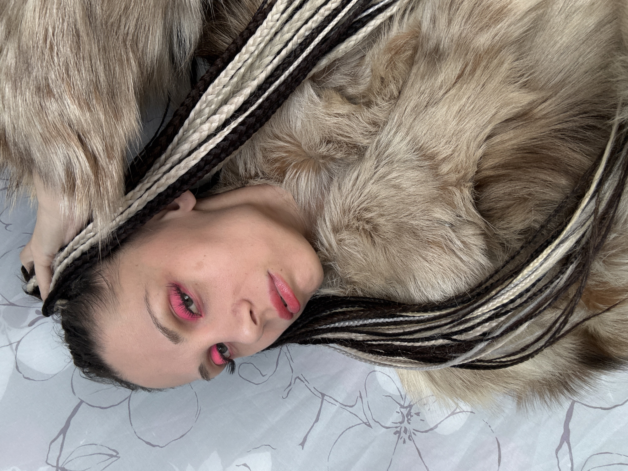 A person is lying down in an indoor setting. The individual has braids in their hair, with alternating black and white strands. The person is wearing pink makeup on their eyelids and lips. They appear to be positioned on a bed with a white patterned sheet. The individual is covered in fur, likely a fur coat or blanket. The location is not identifiable as Rusanovo, Russia based on the provided image.