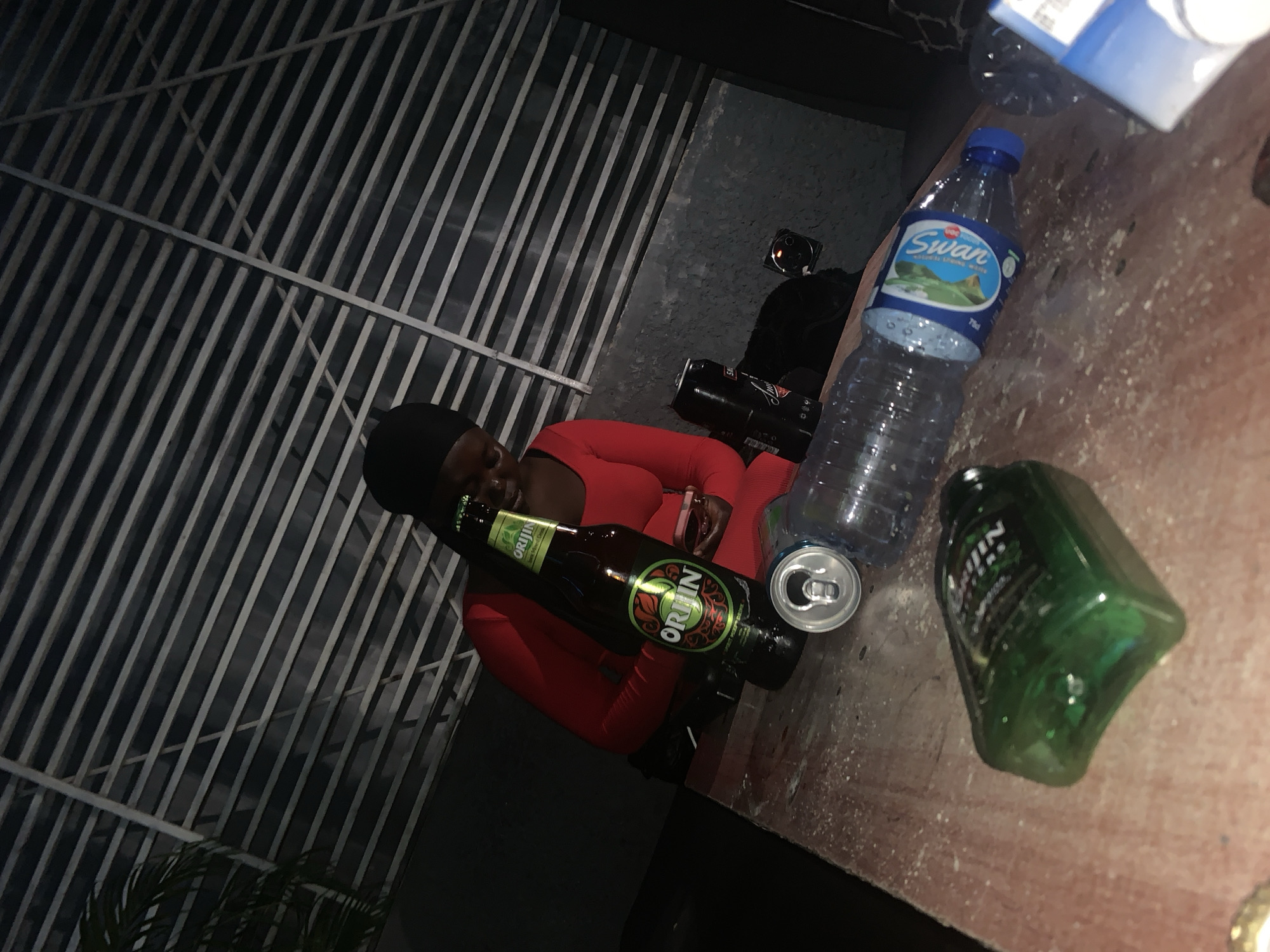 A person wearing a red dress and a black headscarf is sitting at a wooden table outdoors, possibly at night. On the table, there are several objects, including a green glass bottle of Orijin, a can, a plastic water bottle labeled "Swan", and another green bottle. In the background, there is a slatted structure, and the overall environment appears to be urban, consistent with a location in Jos, Nigeria. The lighting is dim, suggesting it is evening or night.