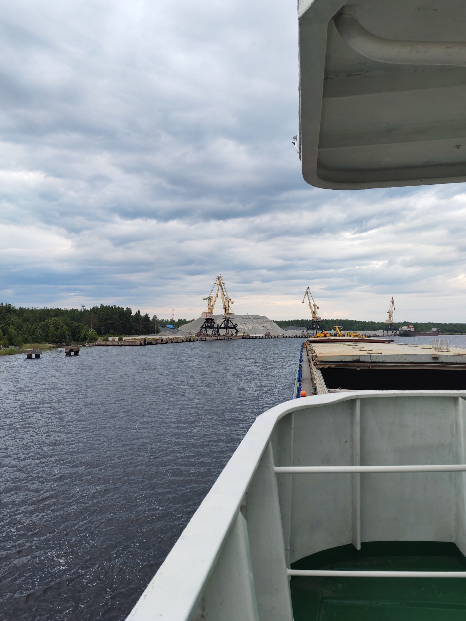 The image depicts a cloudy day at a port, likely in Shal'skii, Russia. The viewpoint is from the deck of a ship looking towards a shoreline. In the foreground, the white railing and a portion of the deck of the ship are visible. The water is dark and somewhat choppy.
In the middle ground, a pier is visible, lined with concrete pontoons. Piles of gravel or sand are stacked on the pier, and several large yellow cranes are positioned to load or unload the material. Some smaller yellow construction vehicles are also present on the pier. Further in the distance, a forested shoreline stretches across the horizon.
On the water, a few small boats are anchored or moored near the shoreline. A larger cargo ship is visible in the far distance, beyond the pier and cranes. The sky is overcast with heavy, gray clouds. The overall scene suggests industrial activity at a port on a cloudy day.