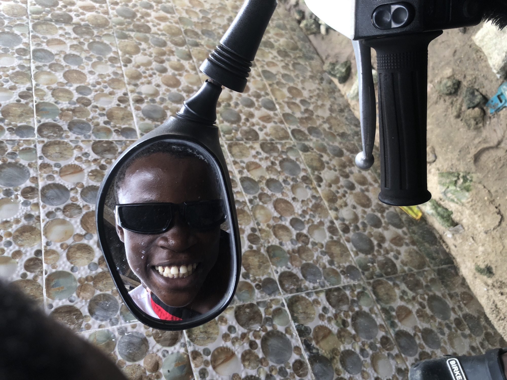 A close-up, low-angle shot captures a person's face reflected in a motorcycle mirror, in Ajah, Nigeria. The person, wearing dark sunglasses, smiles broadly, revealing their teeth. Their head is tilted, and the reflection shows their forehead, eyes, nose, mouth, and chin. The background behind the mirror is a tiled wall with a pattern of circular, stone-like designs in various shades of brown, beige, and gray. In the foreground, the black handlebar of the motorcycle is visible, along with a silver brake lever and part of the white front of the motorcycle. The ground appears to be sand with some scattered rocks and a small, blue plastic object.
