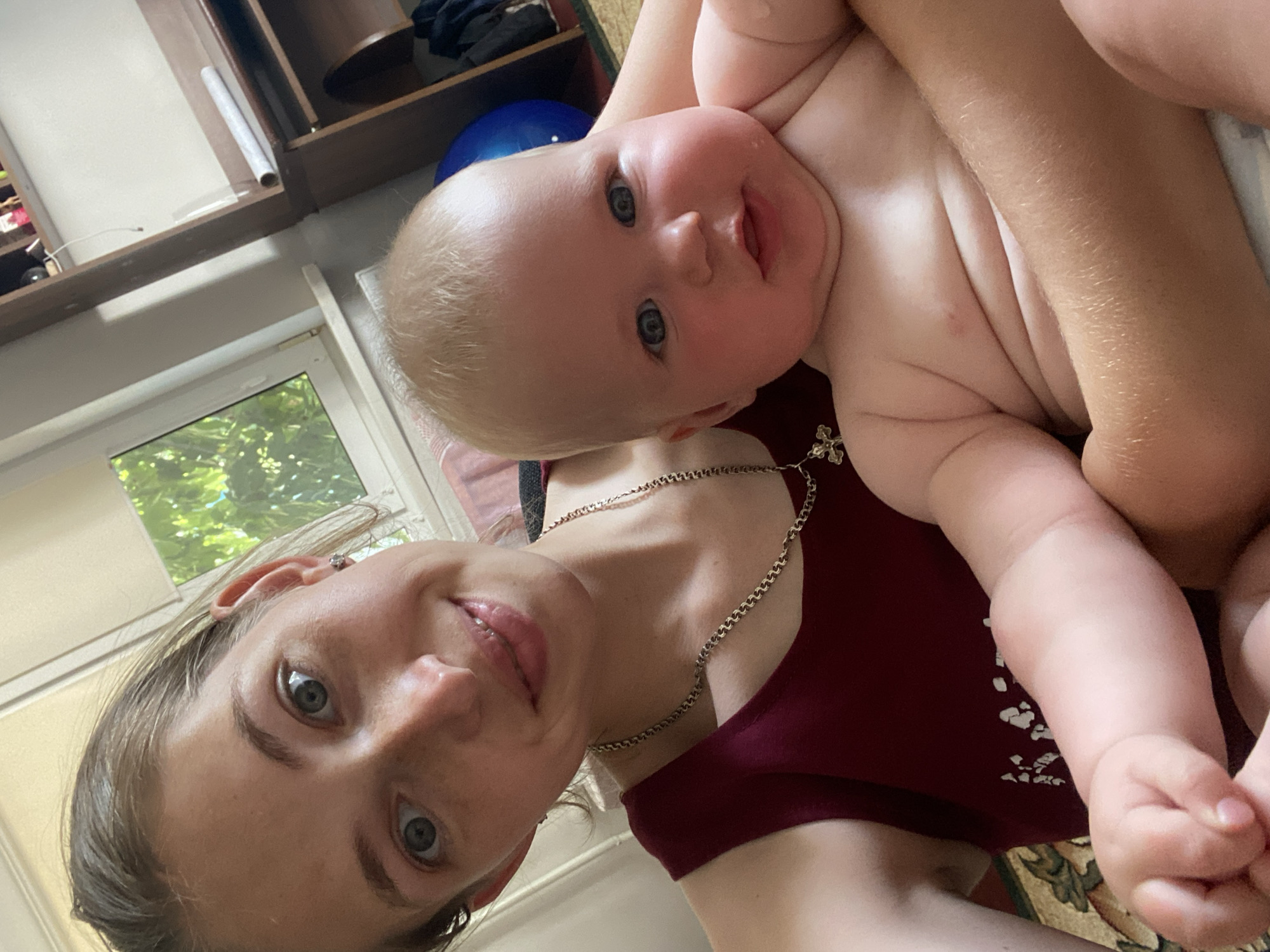 The image shows a woman and a baby. The woman is in the foreground, with her face tilted upwards and towards the camera. She has light brown hair and blue eyes, and is wearing a dark red top and a silver chain necklace. The baby is held in her arms, with its head resting on the woman's shoulder. The baby has blond hair and blue eyes, and is looking at the camera with a slight smile. The background is somewhat blurry, but appears to be an indoor setting with a window showing greenery outside. There are shelves with objects on them in the upper left corner. The lighting suggests it is daytime. The overall mood of the image is warm and intimate, capturing a moment between a mother and child.