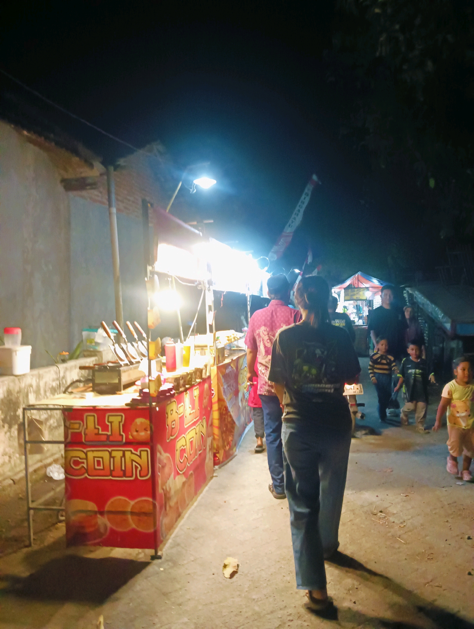 In the city of Unknown, at night, a food stall with a red banner displaying "B-LI COIN" is illuminated by bright lights. Several individuals are walking along a paved pathway beside the stall. A woman in a dark t-shirt and blue jeans is in the foreground, walking away from the camera. Behind her, a person in a pink shirt is visible. Further down the path, a group of people, including children, are walking. The environment appears to be an outdoor market or street setting with a building wall on the left and trees and other stalls in the background.