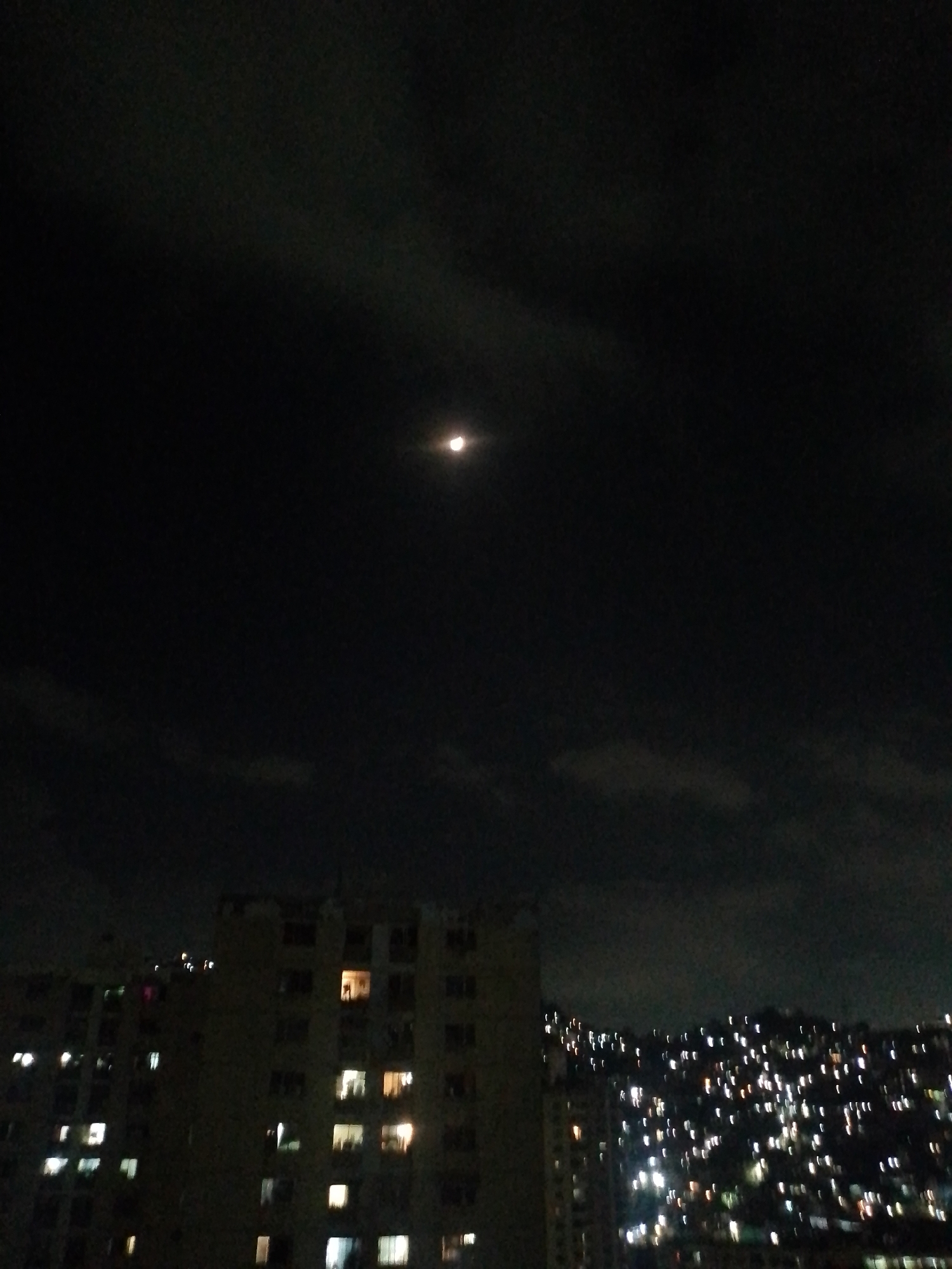 The image depicts a night view in Caracas, Venezuela. A full moon shines dimly in the sky above several apartment buildings, one of which is the primary focus and has several illuminated windows. In the background, lights from other buildings are visible on a hillside. The sky is dark and partly cloudy. No people are visible.
