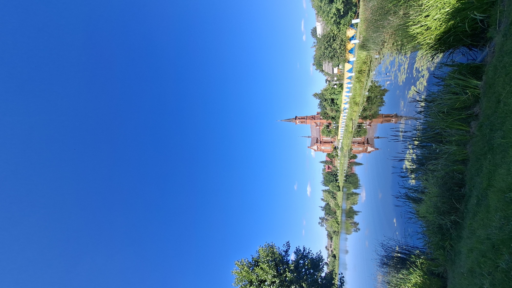 The image depicts a serene scene in Pastavy, Belarus, featuring a large, red brick church with a prominent steeple, its reflection mirrored in the calm waters of a nearby lake. The sky above is a vibrant, cloudless blue. Lush green trees and foliage line the banks of the lake, framing the church and the surrounding landscape. In the foreground, tall grasses and reeds grow along the water's edge, and a patch of green lawn is visible. A decorative fence, with a pattern of yellow and blue triangles, runs along the bank in front of the church. The overall impression is one of tranquility and natural beauty, with the architectural landmark of the church adding a touch of historical character to the picturesque setting.