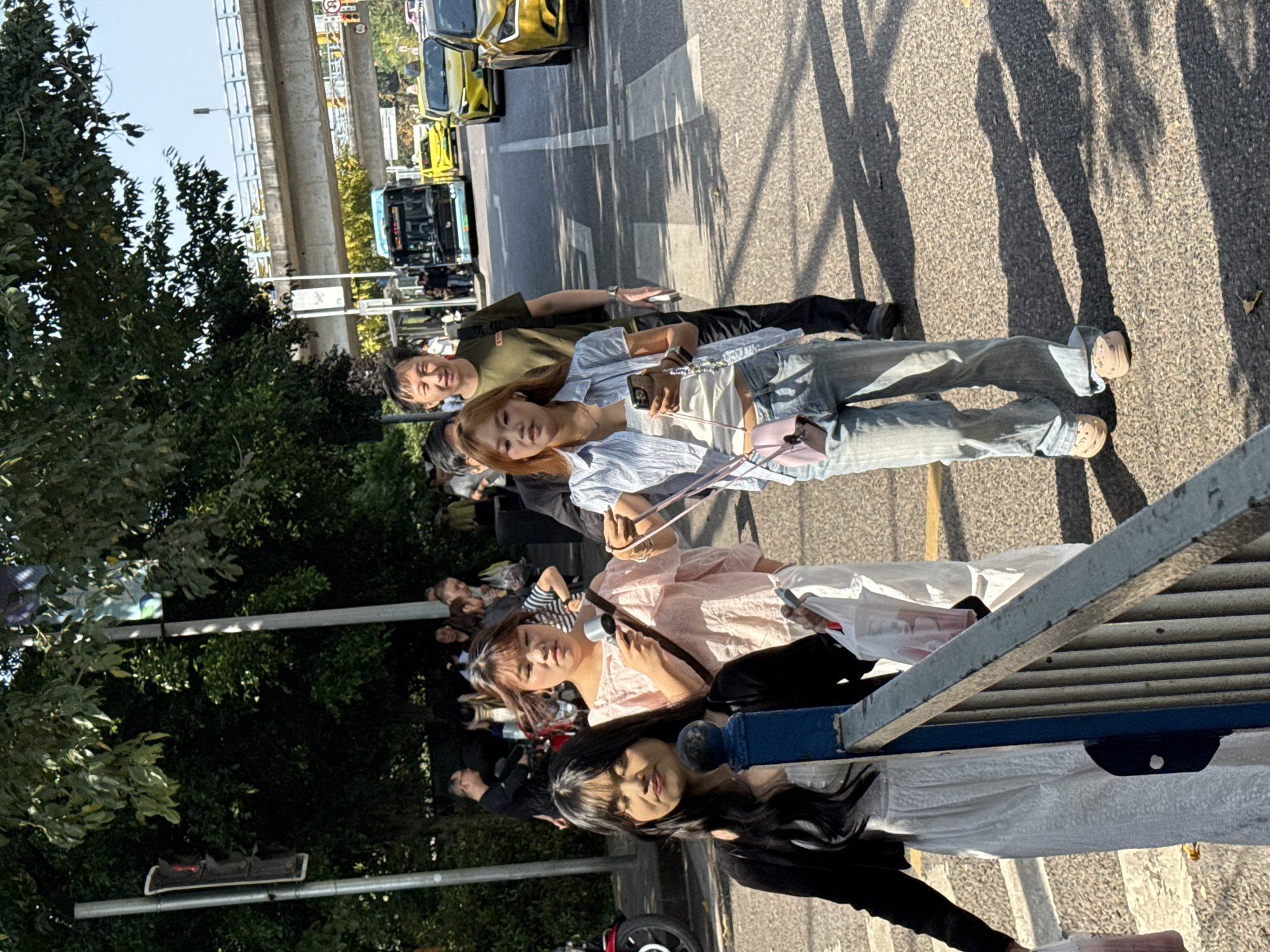 The image captures a vibrant daytime street scene in what appears to be an urban area, likely Nan'an District, China. The bright sun casts strong, distinct shadows on the paved ground, indicating clear, sunny weather.
In the foreground, a metal barrier runs diagonally across the bottom part of the frame, partially obscuring some individuals. Prominently featured are two young women. One, with long dark hair, is visible behind the barrier, looking generally towards the right. The other, with lighter brown hair and wearing a light pink, possibly ruffled, blouse, stands slightly further back, holding a silver object that could be a microphone, a handheld fan, or a small camera accessory. She is also looking towards the right, appearing engaged with something beyond the frame.
Behind these individuals, a group of people is gathered. A man in a dark green shirt and black pants stands with a relaxed posture, looking forward, while a woman next to him in a light blue striped shirt and jeans holds a smartphone. Numerous other individuals are visible in the mid-ground and background, suggesting a crowd or a public gathering.
To the left of the scene, lush green trees and bushes line the street. On the road itself, several yellow cars, possibly taxis, are visible, along with a blue public bus. An elevated concrete road or overpass runs parallel to the street in the distance. The overall atmosphere suggests a casual outdoor event or a busy public area. Although some signs are visible on the elevated structure and along the road, any text on them is illegible.