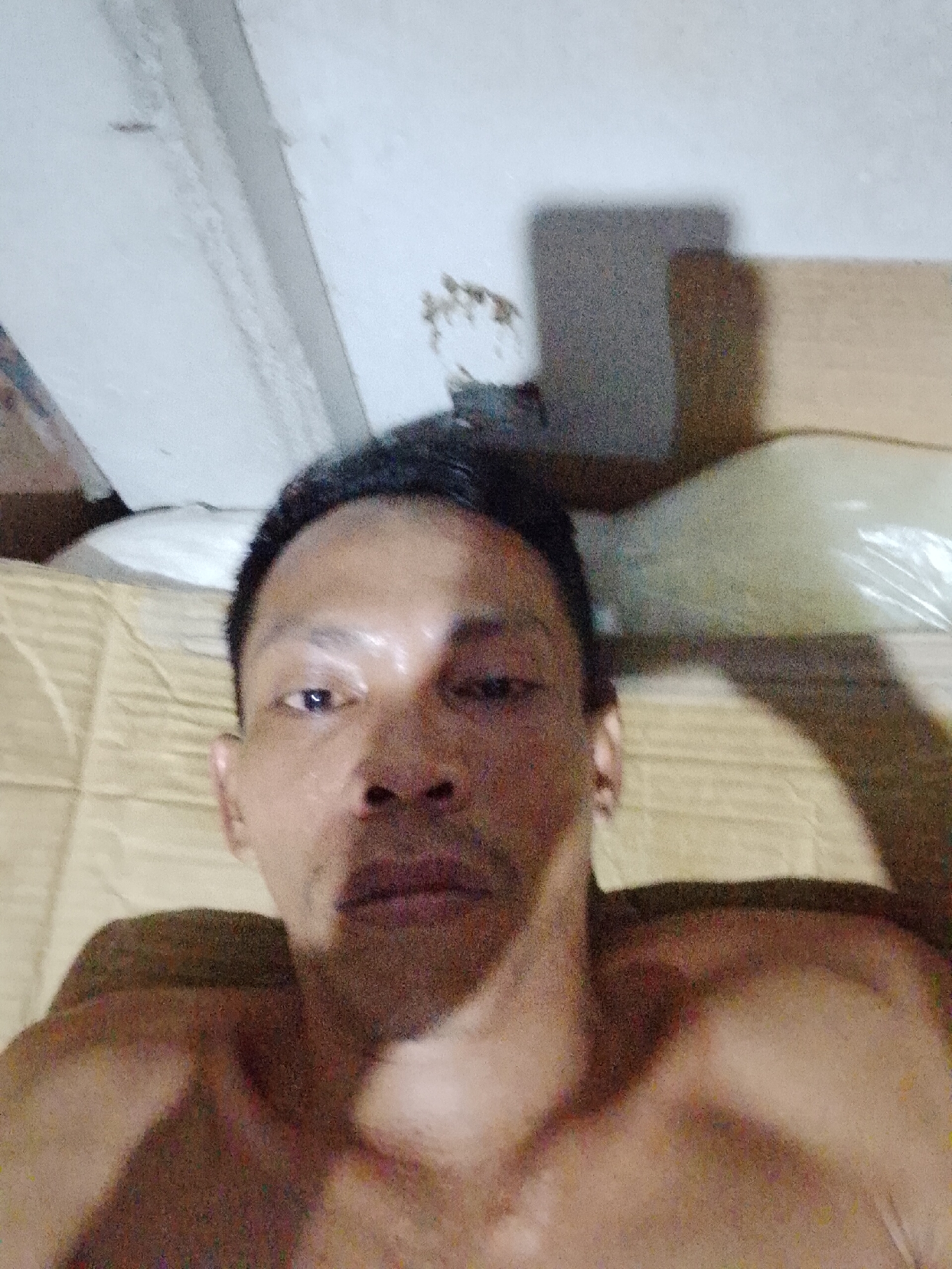 A shirtless person is lying down, presumably on a cardboard surface. A ceiling is visible in the background, along with some boxes. The location is Bukit Ketok, Indonesia, based on the prompt.