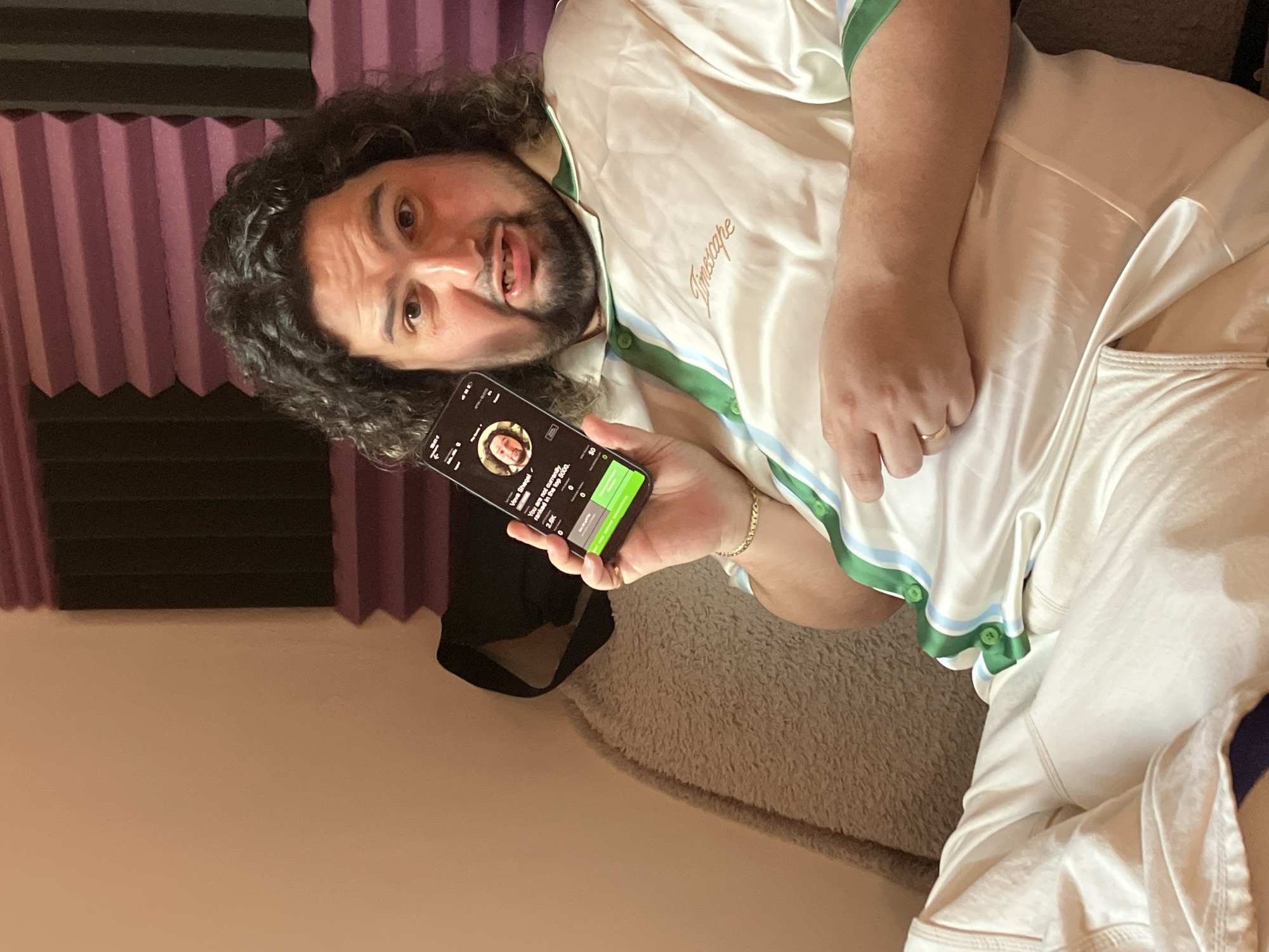 A man with dark curly hair, a beard, and a white silk pajama shirt with green trim is lying on a neutral-colored textured surface, possibly a couch or bed. He is holding a smartphone showing a music app, and he is looking directly at the camera with a slight expression of surprise. Behind him is a wall with purple soundproofing panels. The exact location in Tel-Aviv, Israel cannot be determined from the image.