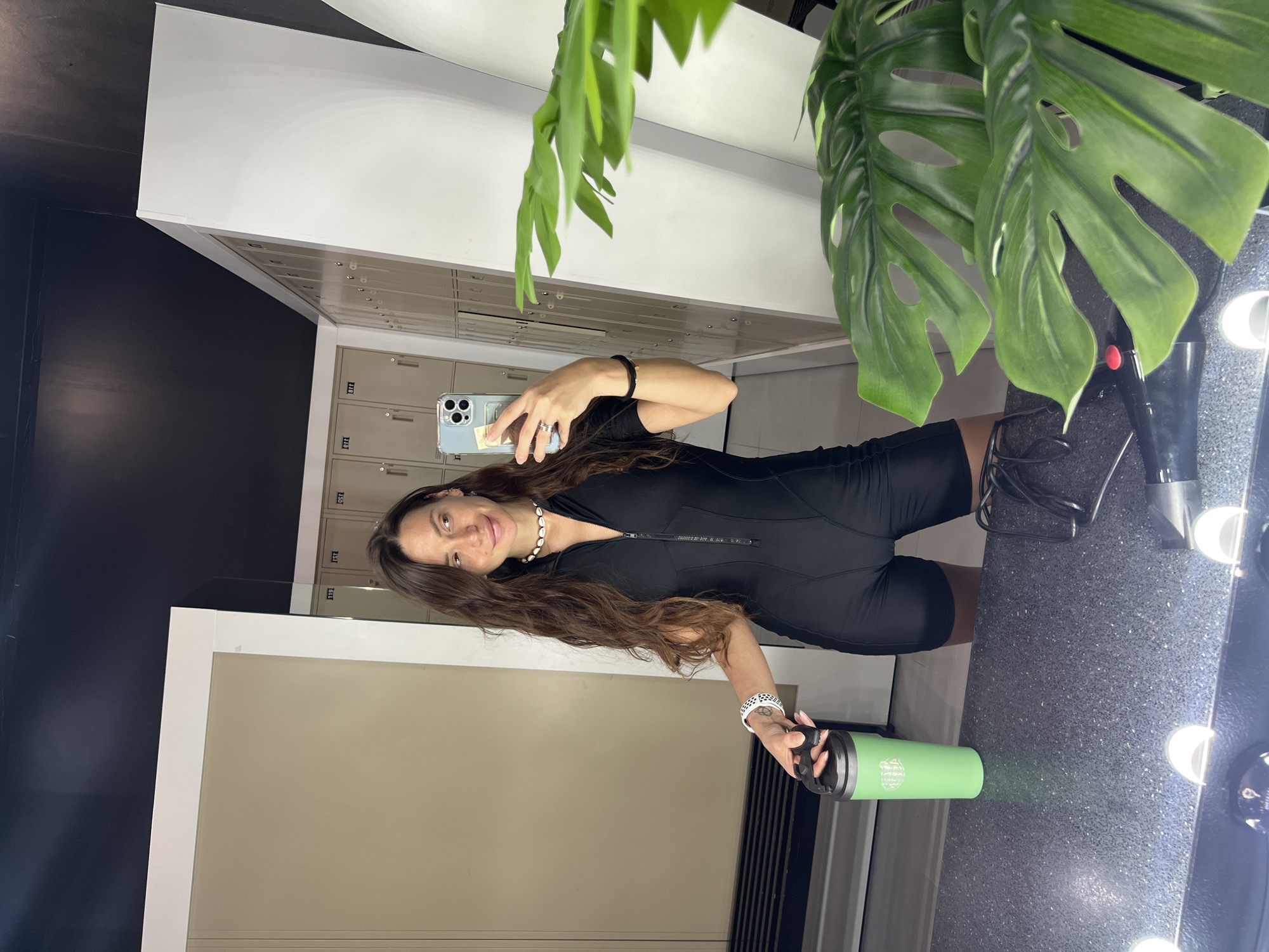 A person is taking a selfie in a locker room. The person is holding a green water bottle in their left hand and a smartphone in their right hand. The person is wearing a black bodysuit, a shell necklace, and a white smartwatch. The person has long brown hair. The background includes lockers, a mirror with vanity lights, and a potted plant. The location is Unknown, Unknown.