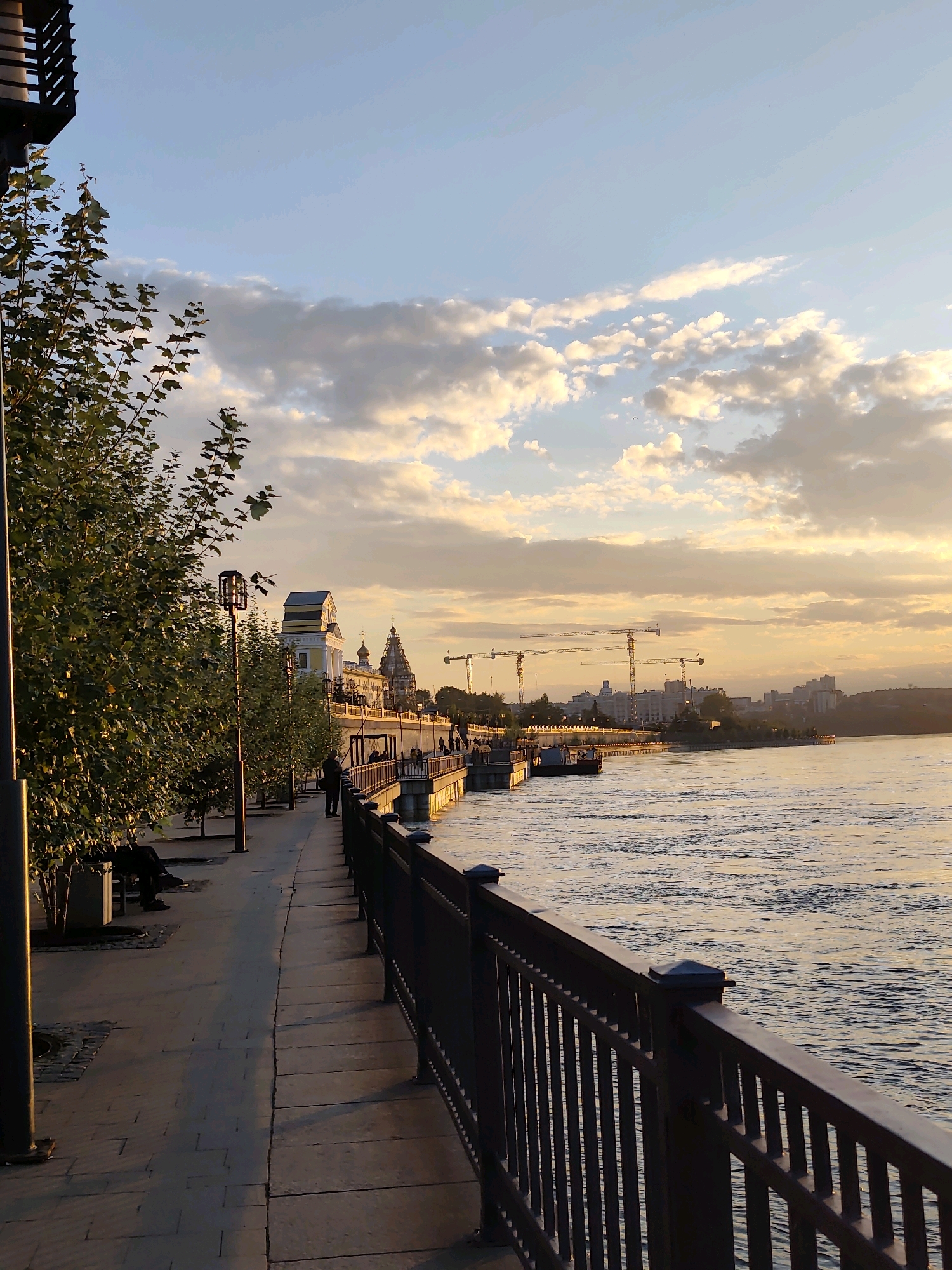 The image captures a scenic view of the Angara River embankment in Irkutsk, Russia, during what appears to be late afternoon or early evening. The setting sun casts a warm, golden glow across the water and the sky, illuminating scattered, fluffy clouds.
On the left side of the frame, a paved pedestrian walkway runs alongside the river. It is lined with trees, some of which are fully leafed, and streetlights. A few individuals are seated on benches, and one person is walking along the path. The walkway is bordered by a dark metal railing, which separates it from the river.
Across the river, a cityscape is visible, featuring buildings, including what appears to be a church with multiple domes and a lighter colored building with a distinctive roof. Several construction cranes are also present in the background, suggesting ongoing development. The river itself is calm, with gentle ripples reflecting the sky's colors. The overall atmosphere is tranquil and picturesque.