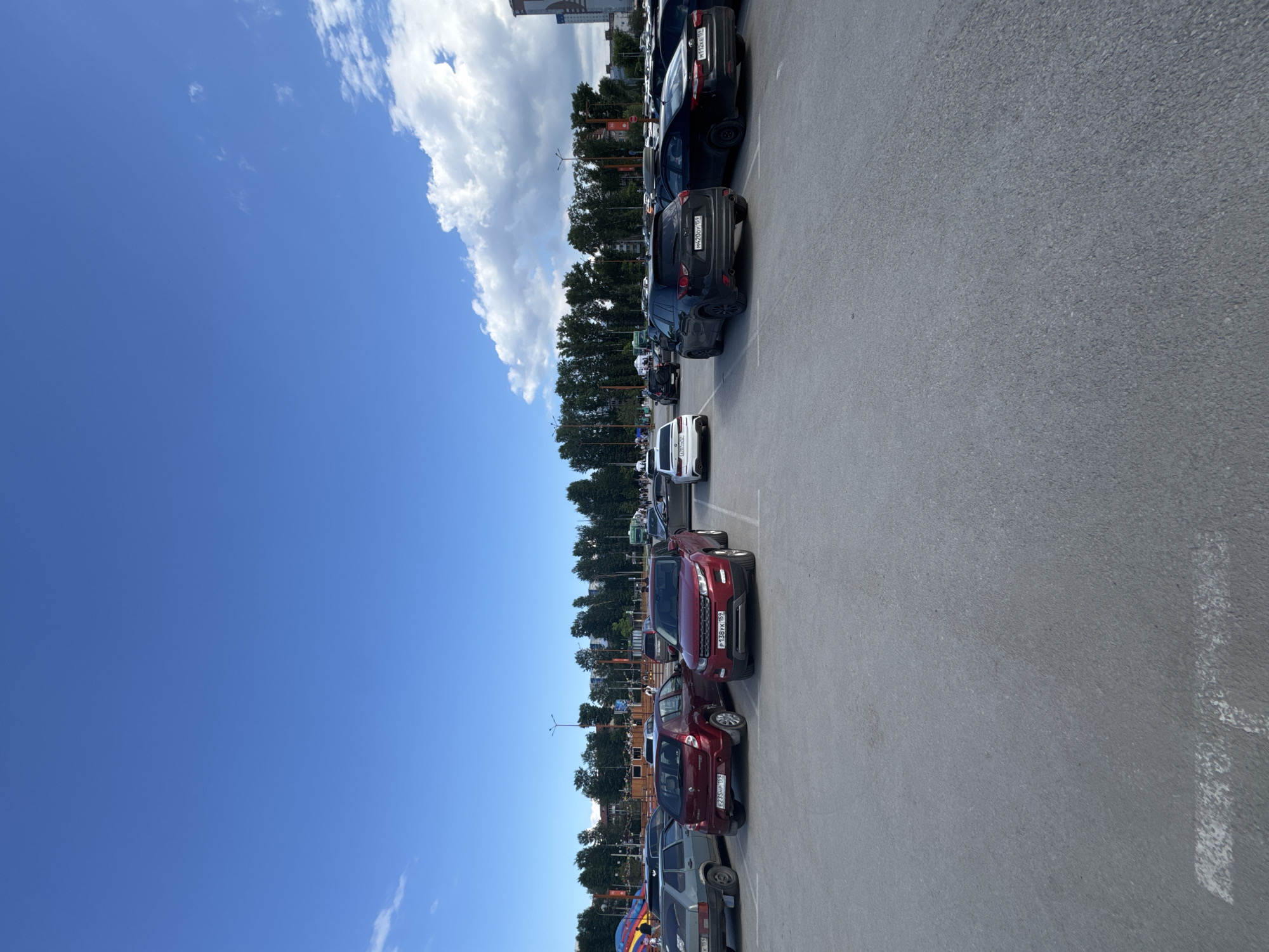 The image shows a parking lot in Perm, Russia. Several cars are parked in a row, including a red Range Rover Evoke, a white car, and other vehicles in various colors. The parking lot appears to be located near a building and is surrounded by trees. The sky is blue with some clouds.