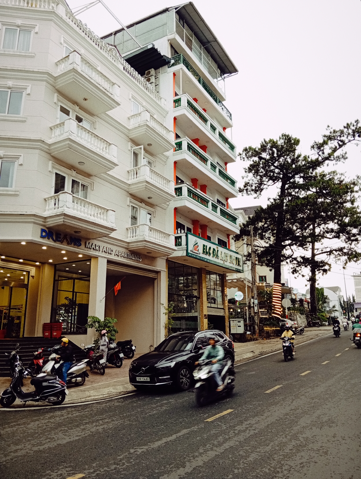 The image captures a lively street scene in Da Lat, Vietnam, characterized by multi-story buildings, numerous motorbikes, and prominent trees under an overcast sky. On the left, a grand white building with ornate balconies and white railings prominently displays "DREAMS MART AND APARTMENTS" on its ground-floor signage. Next to it, another building features balconies with green railings and red support pillars, identified as "ELC DA LAT HOTEL" with its address "ĐC: 23 BÀ TRIỆU P2 TP. ĐÀ LẠT."
The wet asphalt road, possibly from recent rain, is marked with yellow lines. Numerous motorbikes are parked along the sidewalk, and several are in motion on the road, with some riders blurred from movement. A black SUV bearing license plate "51K-134.61" is parked curbside. Individuals are visible on or near their motorbikes, contributing to the bustling urban atmosphere.
Large trees with dark green foliage line the right side of the street; one tree trunk is wrapped with decorative string lights, suggesting a festive mood or evening setting. A Vietnamese flag hangs near the entrance of the "Dreams Mart" building. The light grey, overcast sky provides muted illumination, indicating either a cloudy day or late afternoon/early evening in this busy Vietnamese city.
