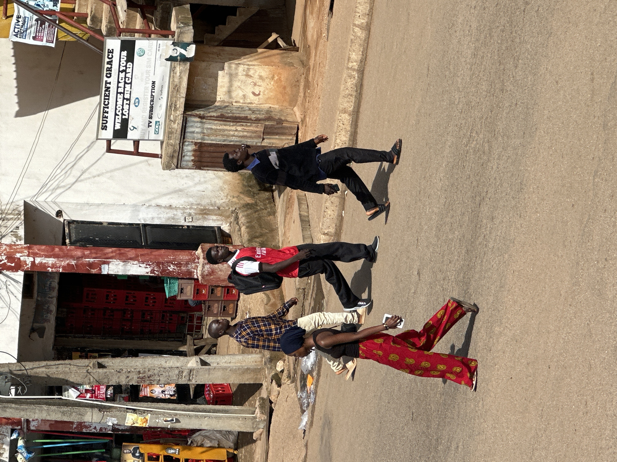 In Bukuru, Nigeria, four individuals are captured mid-stride on a paved street. A woman in the foreground walks from left to right, dressed in a vibrant red, yellow, and green patterned skirt and a black sleeveless top, holding a white object in her right hand. Behind her, a man in a plaid shirt and light-colored pants walks from left to right, his left hand in his pocket. To his right, a man in a red t-shirt and black pants, carrying a black backpack, walks from left to right, looking forward. Further to the right, a man in a black sweater and black pants, holding a dark object in his right hand, walks from left to right. The background features buildings with faded paint, some displaying advertising signs, and stacked red crates, suggesting a commercial area. Several concrete utility poles line the left side of the street, with wires running between them. The street is mostly clear, with some scattered litter. The lighting indicates it is daytime, with shadows cast on the ground.