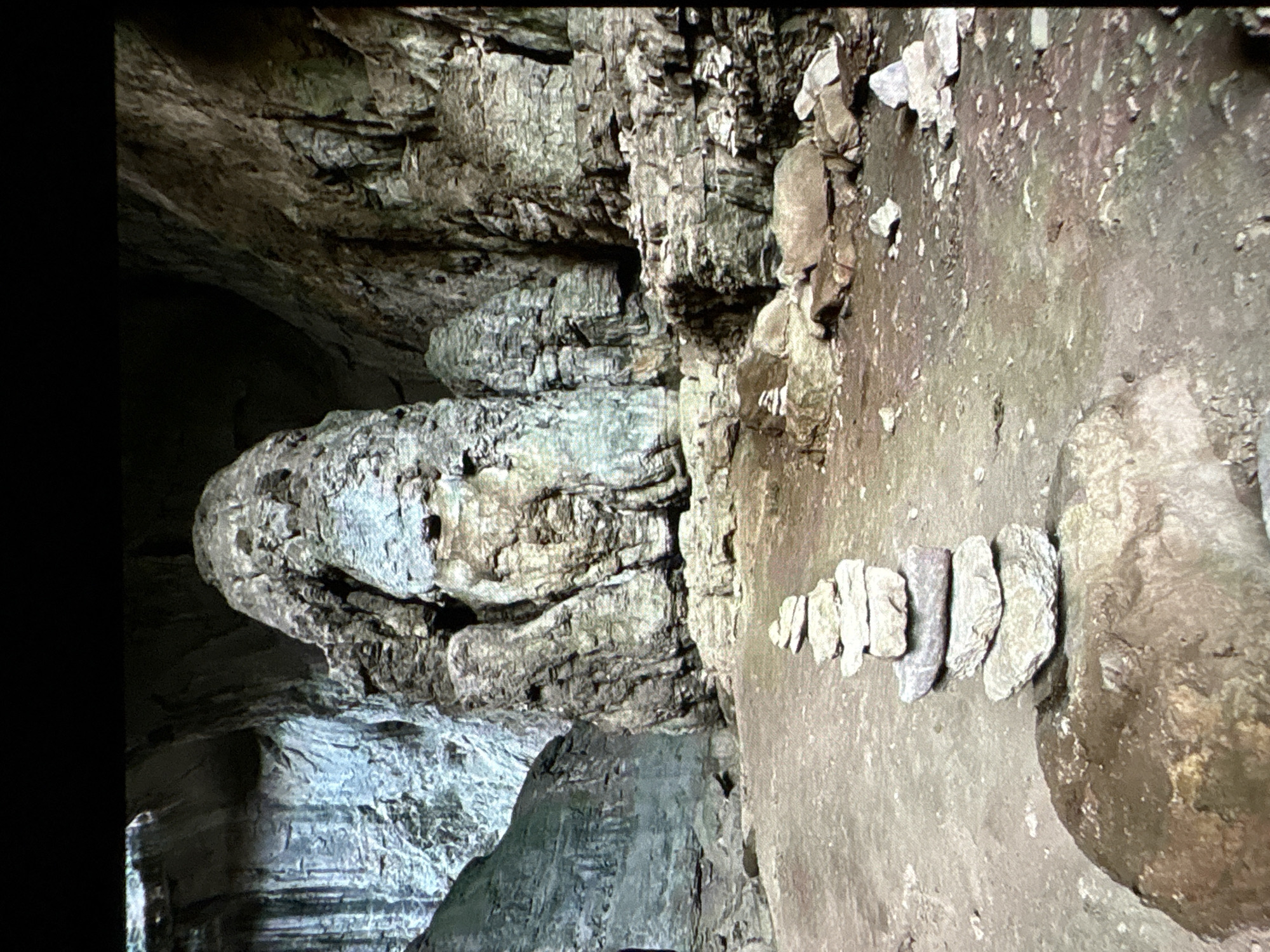 The image shows the interior of a cave. The scene is dominated by rough, textured rock formations, with layers and crevices suggesting geological processes over time. To the right of the frame, a wall of earth and stone is visible, with a line of stacked flat stones arranged vertically. These stones are of varying sizes and shades of grey and white, creating a deliberate pattern on the earthen surface.
In the center-left of the image, a large, bulbous rock formation juts out from the cave wall, its surface heavily textured and weathered. Behind and around this formation, the cave recedes into darkness, with glimpses of more layered rock. The lighting appears natural, likely from an opening within the cave, casting shadows and highlighting the rugged contours of the rock. The overall impression is one of a natural, undisturbed environment, with the stacked stones being the only clear indication of human presence or activity. The location context of Plovdiv, Bulgaria, does not provide specific identifiable landmarks within the cave itself. There is no visible text in the image.
