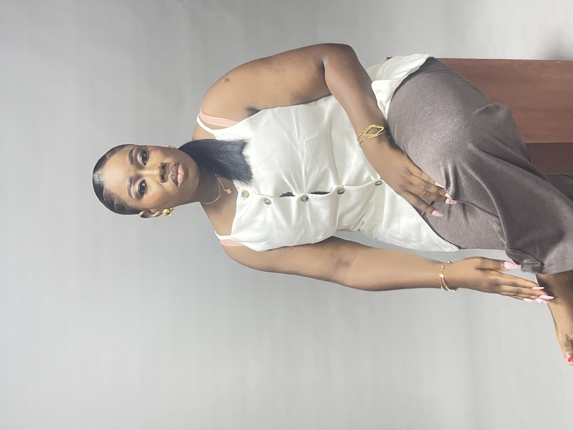 A young Black woman is depicted in a medium shot, reclining or sitting with a plain light gray background. She is looking directly at the camera with a neutral to slightly serious expression. Her dark hair is pulled back, showing sculpted edges, and she wears subtle eye makeup and lip color.
She is dressed in a sleeveless white top with buttons down the front, paired with brown or gray wide-leg pants. Her outfit is accessorized with gold jewelry, including hoop-style earrings, a delicate necklace with a small pendant, and two distinct gold bracelets—one on each wrist. Her fingernails are painted with light pink polish, and her toenails have a reddish hue.
Her right hand rests on her upper thigh, while her left arm is extended forward. Behind her, a brown surface, possibly a wooden bench or chair armrest, is visible. The lighting is even, suggesting an indoor setting. There is no visible text in the image.