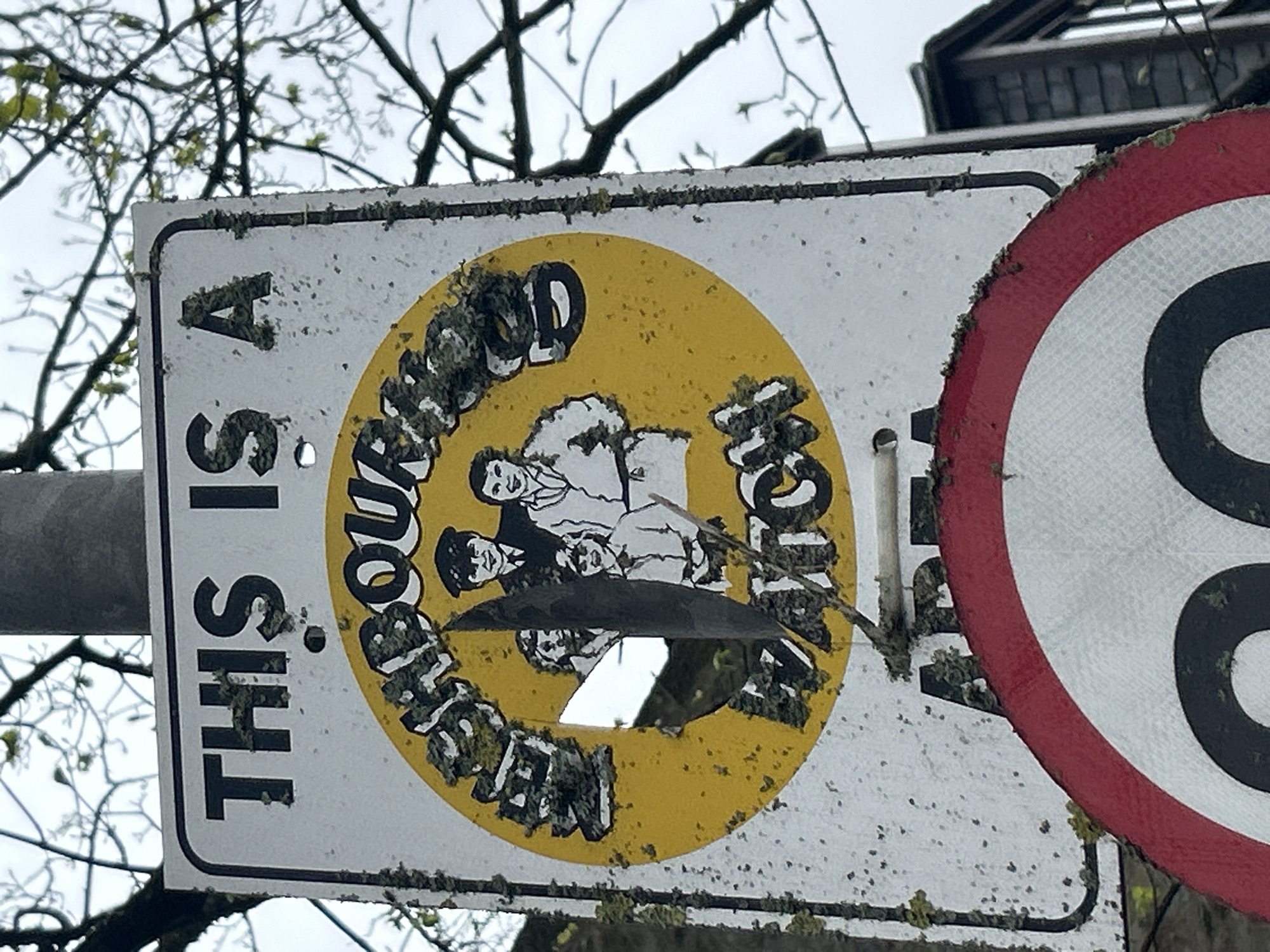The image shows two adjacent road signs in Aberdeen City, United Kingdom. The sign on the left states, "THIS IS A NEIGHBOURHOOD WATCH" around a yellow circle containing a cartoon-style image of a group of people. The sign is visibly aged and has a hole in the center. Lichen and other biological growth are present on the signs. The sign on the right, partly visible, is a circular road sign with a red border, partially obscuring its content. Bare tree branches are visible in the background, suggesting a possible outdoor setting. The sky appears overcast. The entire scene appears to be situated outdoors in a natural environment.