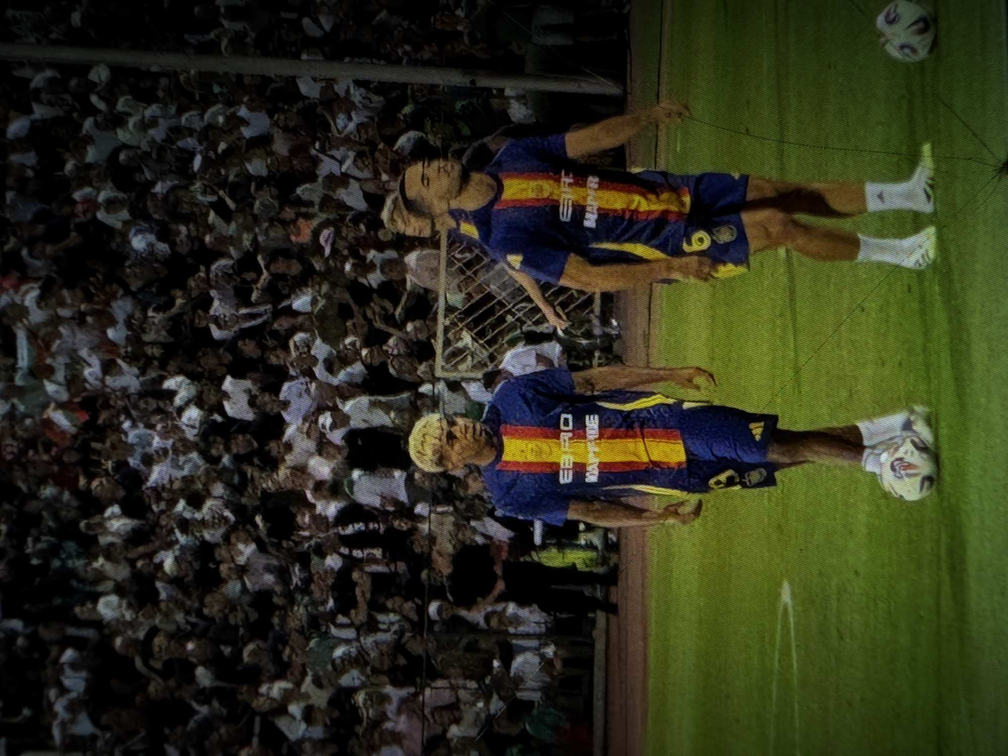 Two male soccer players in blue and red striped uniforms are on a green soccer field with a crowd of spectators in the background. The player on the left has blonde hair and is looking to the right. He is wearing a blue jersey with red and yellow stripes down the middle and blue shorts. The player on the right is looking forward and is wearing a similar uniform. He has the number 6 on his shorts. There is a soccer ball at the feet of the player on the left. The spectators are blurred and appear to be sitting in stands. The scene appears to be a soccer match.