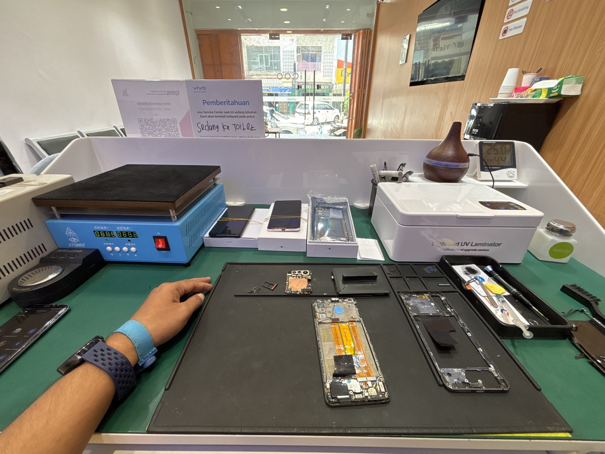 The image depicts the interior of a mobile phone repair shop in Lhokseumawe, Indonesia. A person's left forearm, adorned with a blue anti-static wrist strap and a dark blue smartwatch, is visible in the foreground, resting on a green mat. The mat is covered with disassembled smartphone components, including circuit boards, screens, and internal hardware. Various repair tools, such as screwdrivers and tweezers, are neatly arranged in a black tray. In the background, a blue and silver heat plate machine with a digital display and a red power switch is present, alongside a white ultrasonic cleaner and a UV laminator. A Vivo service center sign with information about temporary closure due to a break is prominently displayed. The shop appears well-lit, with a large glass door in the background offering a view of the street outside, where vehicles and buildings are visible. The overall scene conveys a professional and organized environment for electronic device repair.