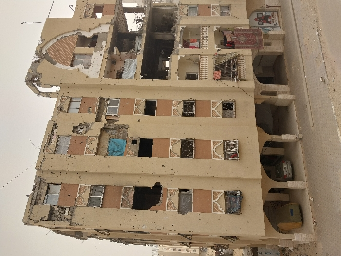 This image displays a five-to-six-story residential or commercial building in Khan Yunis, Palestinian Territory, characterized by extensive structural damage. The beige-painted facade, featuring reddish-brown panels and decorative white patterns around windows, is riddled with numerous small, circular impact marks consistent with bullet holes. Large sections of the exterior walls and interior floors are collapsed or missing, particularly on the upper levels and the right side of the structure, exposing rebar, rubble, and internal rooms. A prominent decorative arch on the upper right side of the building is severely compromised. Most windows are either shattered, absent, or covered by makeshift barriers, including a blue tarpaulin on the third floor (from bottom, left) and black tarpaulins on the second floor (from bottom, left). Damaged balconies on the lower right feature hanging items, including a large red patterned rug and various pieces of fabric. The ground floor consists of open arched bays, providing shelter for a silver sedan and a yellowish-green covered object on the left. The foreground shows a paved road surface with scattered dirt and debris under an overcast, hazy sky. No individuals are visible in the scene.