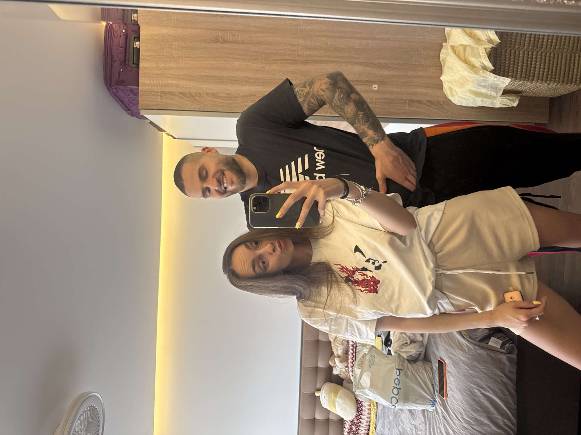 Two people are taking a selfie in a room. A man with a shaved head, beard, and tattoos on his arm is standing next to a woman with long brown hair. The woman is holding a phone, presumably taking the picture. The room appears to be a hotel room based on the luggage and furniture. There is a bed in the background, a closet, and other items scattered around. The exact location in Sofia, Bulgaria, is not discernible from the image.