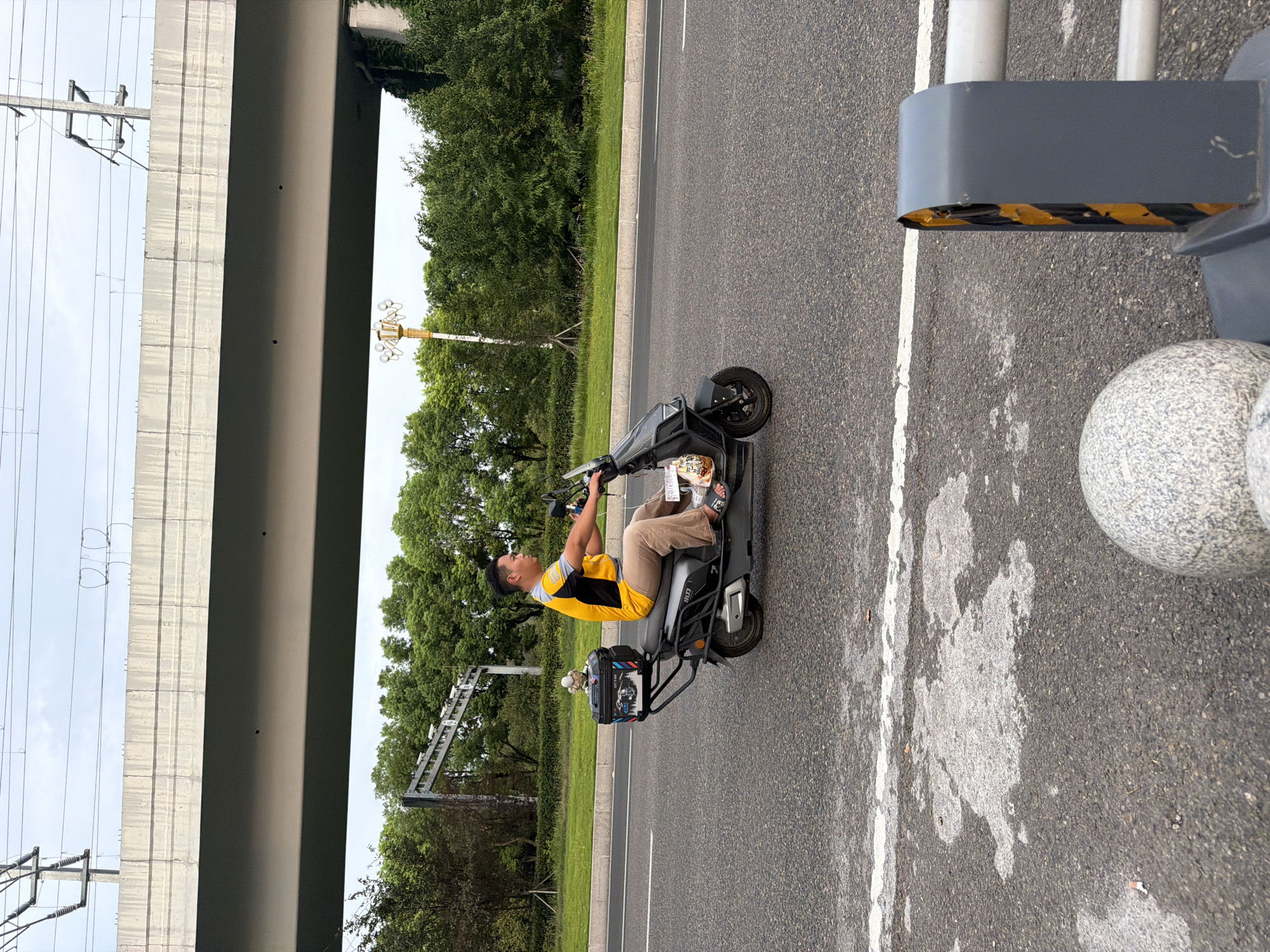A man wearing a yellow and black shirt and khaki pants is riding a dark gray electric scooter on a paved road. He is looking to his right. The scooter has a black storage box on the back with a small figurine on top. The man is wearing sandals. The road has white lane markings. In the background, there are trees and a large concrete overpass with power lines. A lamppost with multiple lights is visible behind the trees. The scene appears to be in an urban or suburban area of Jiangjin, China.