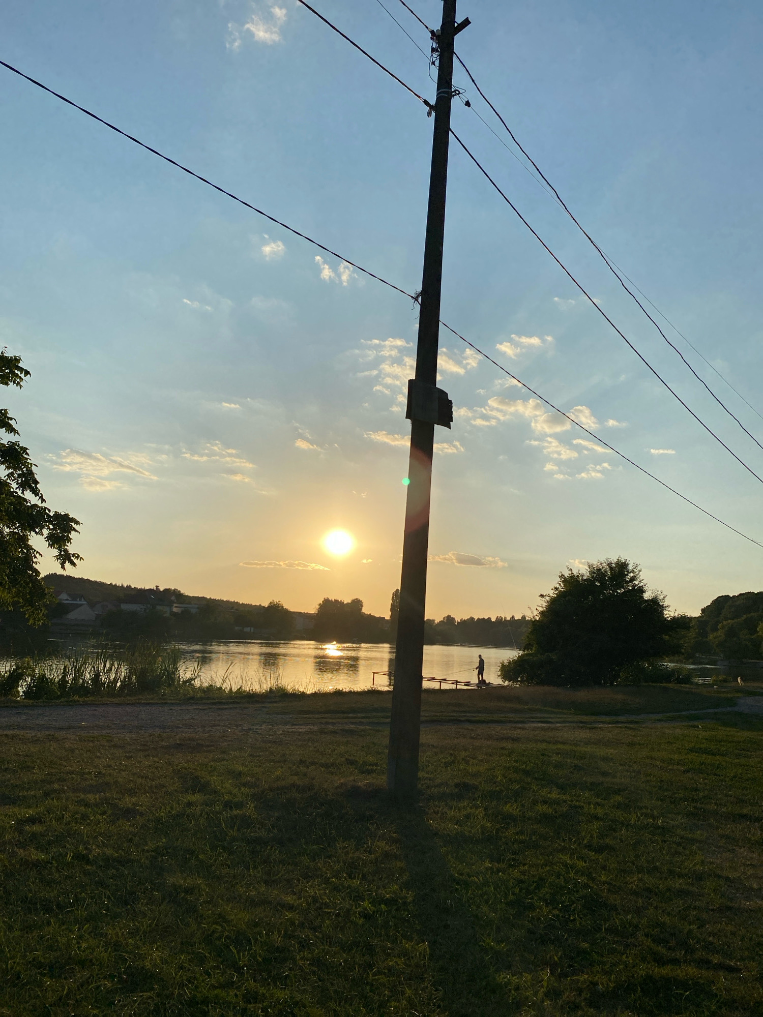 This photograph captures a serene sunset over a body of water, likely a river or lake, in Vinnytsia, Ukraine. The sky is a gradient of pale blue and warm yellow, with scattered white clouds. The sun, a bright orb, is positioned in the middle right of the frame, casting a golden reflection on the water's surface.
In the foreground, a grassy area with scattered shadows suggests late afternoon or early evening light. A weathered utility pole with several wires stretching across the frame dominates the center of the image. Beyond the pole, the water stretches towards a tree-lined shore on the opposite bank, where houses with pitched roofs are visible amongst the trees, indicating a residential area.
On the water's edge, a solitary figure is standing on what appears to be a wooden pier or jetty, holding a fishing rod, suggesting the activity of fishing. Further in the background, several trees are silhouetted against the setting sun. The scene evokes a sense of tranquility and a peaceful rural or suburban atmosphere on the outskirts of the city.